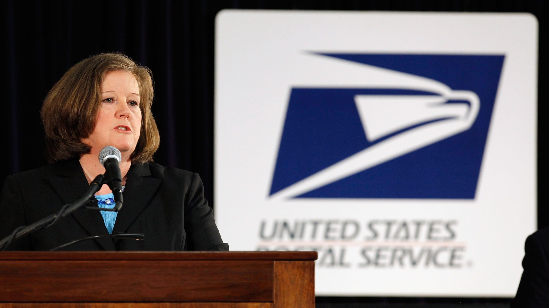 In this image, Brennan speaks from behind a podium while the U.S. Postal Service logo stands behind her.