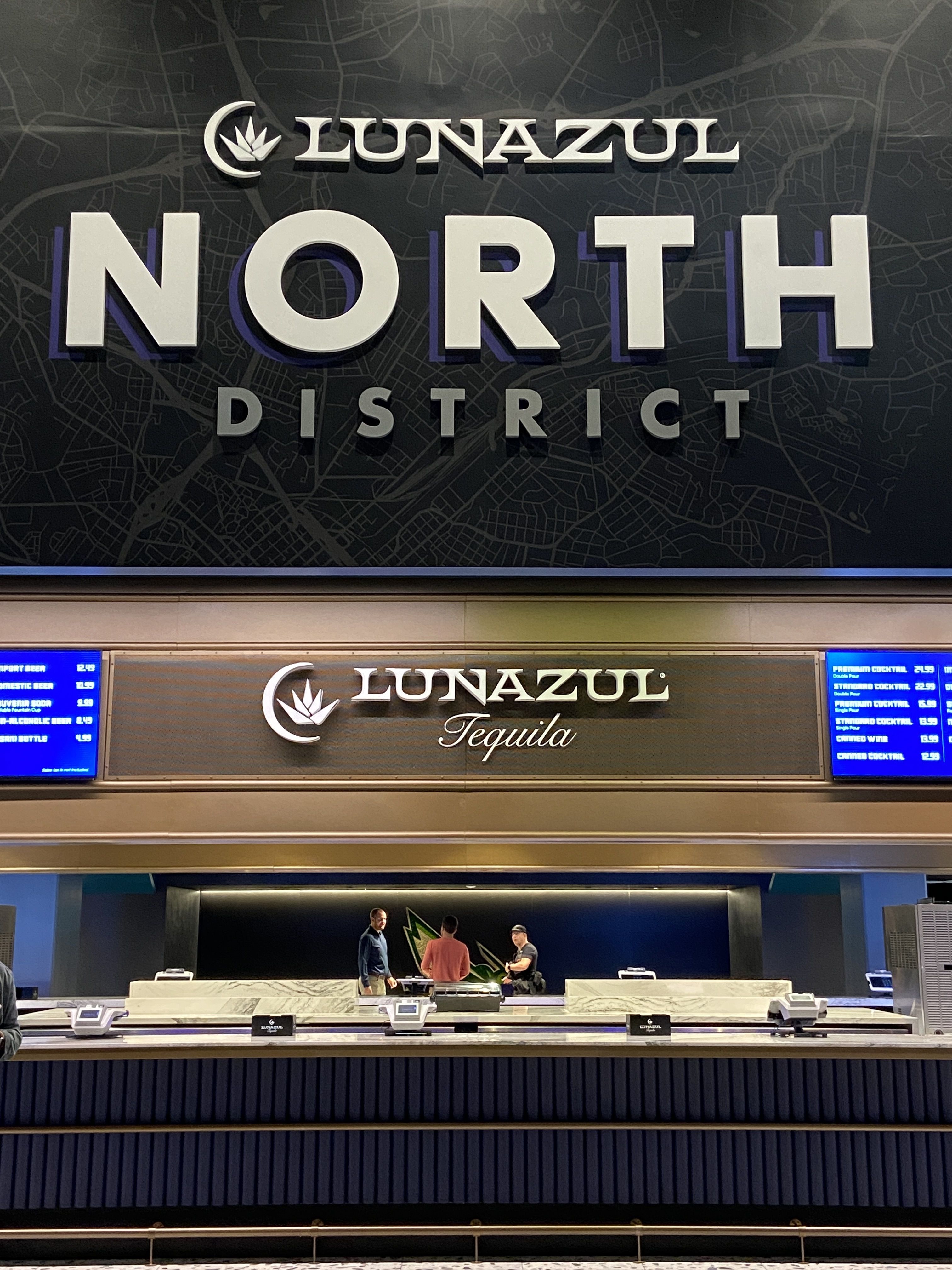 Bar area with large sign reading LUNAZUL NORTH DISTRICT in white on dark background, a smaller LUNAZUL Tequila sign above marble counters, three people behind the bar.