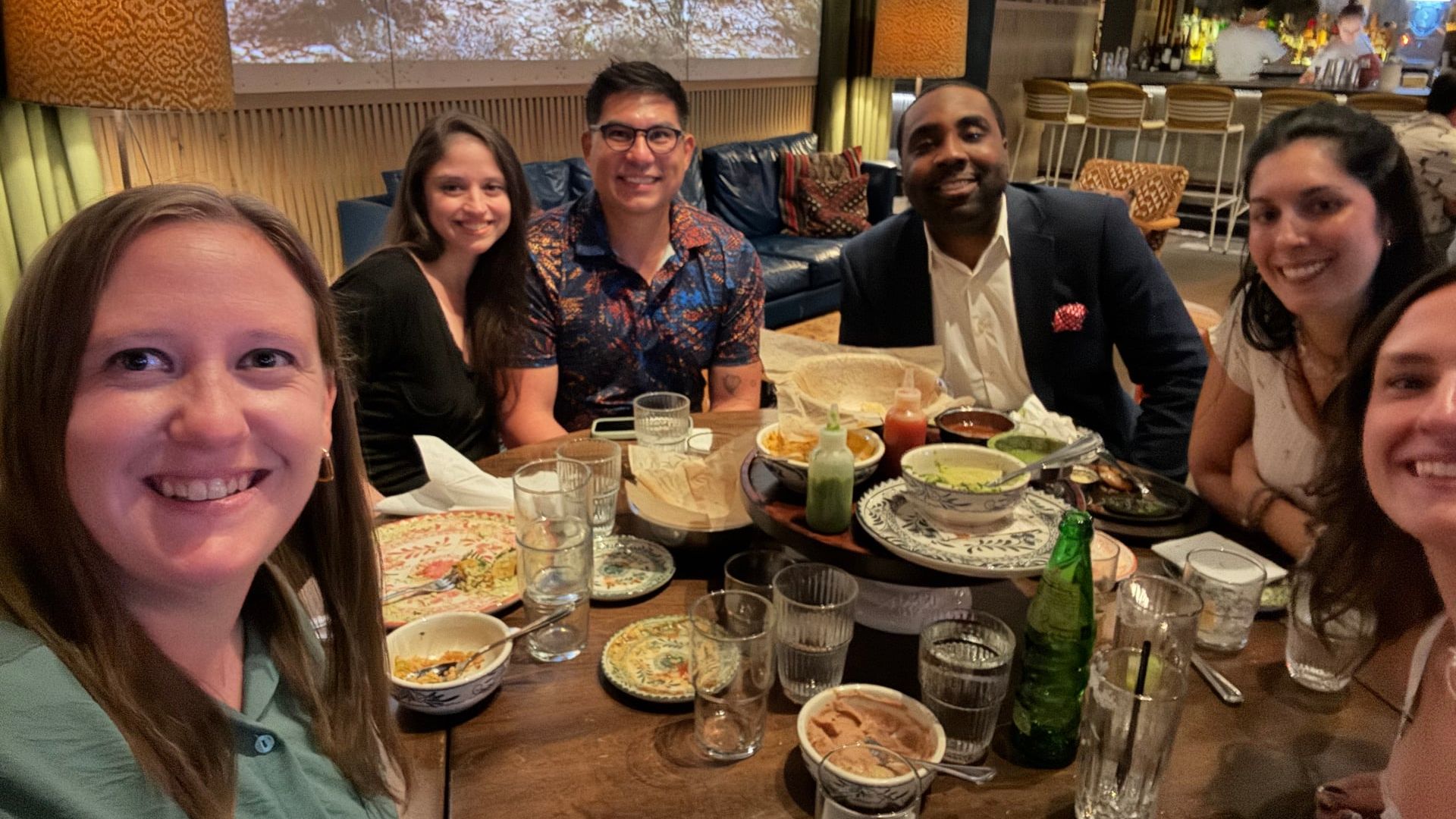 Six friends sit around a wooden table in a cozy restaurant, smiling at the camera. Plates, bowls, sauces and drinks fill the table; warm lighting and a bar backdrop complete the scene.