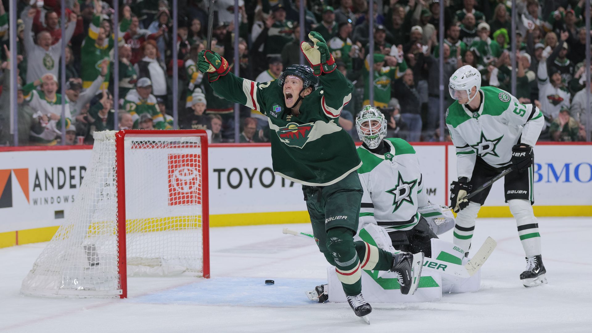 A hockey player in green raises arms in celebration by the net, with cheering crowd behind glass; the goalie kneels and an opponent in white skates nearby, red goal frame visible.