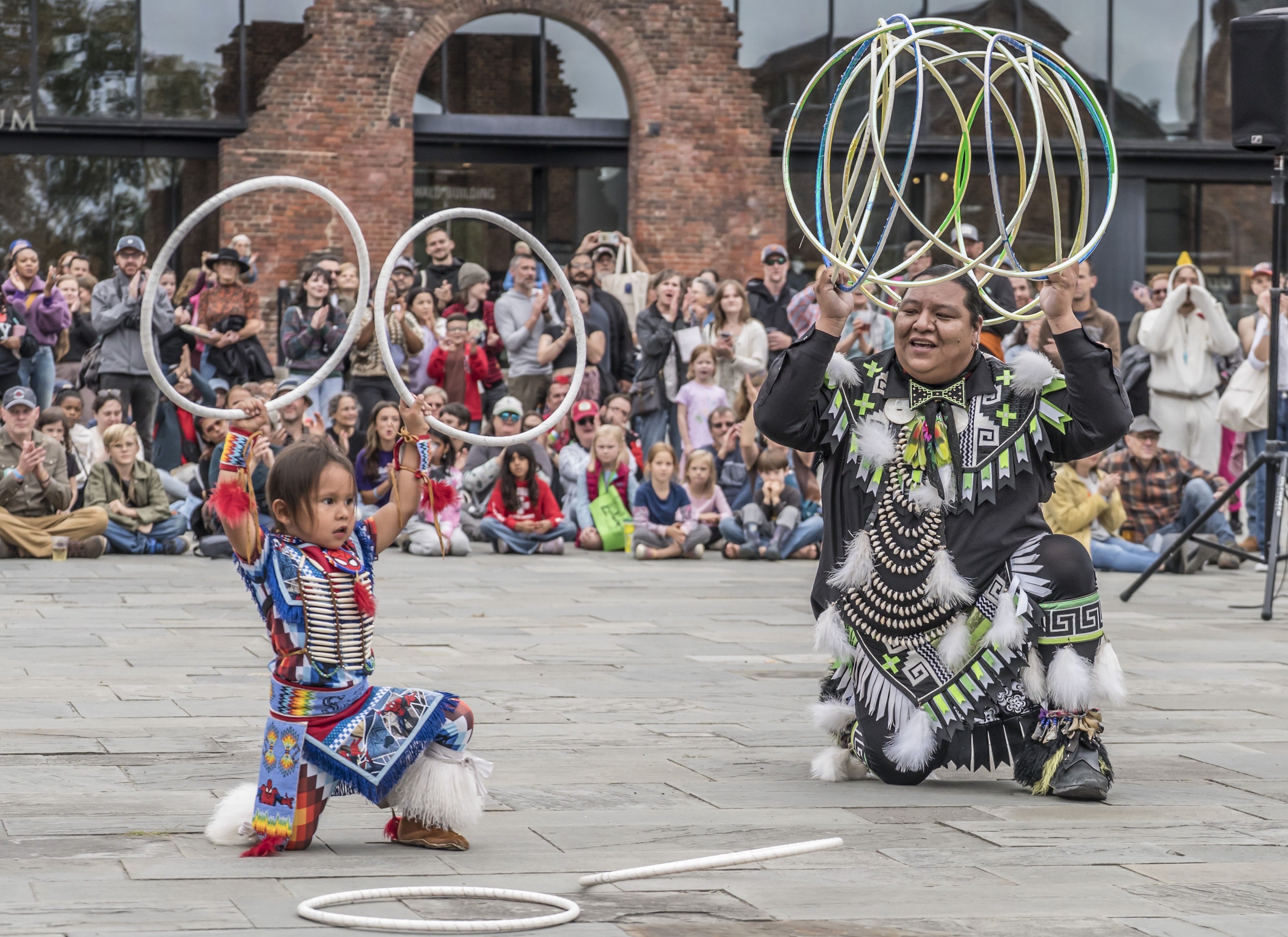 Street performance in a plaza: a dancer in a black geometric costume holds many hoops overhead while a child in bright patterns spins a hoop; crowd watches near brick buildings.