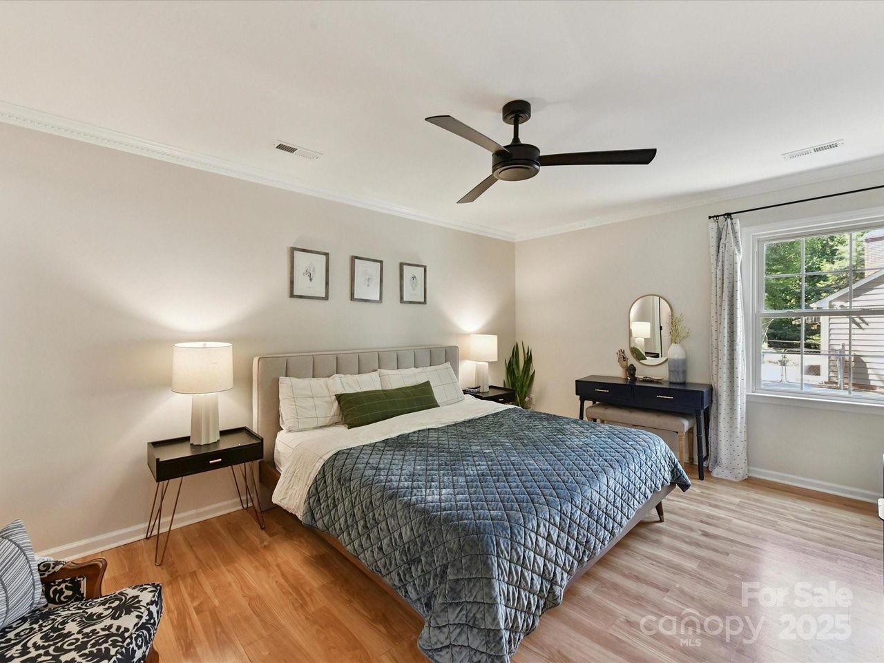 Bright bedroom with a large bed featuring a blue quilt and green pillow, two black nightstands with lamps, three minimalist framed artworks, a black ceiling fan, a window with white curtains, and a black vanity table with mirror.