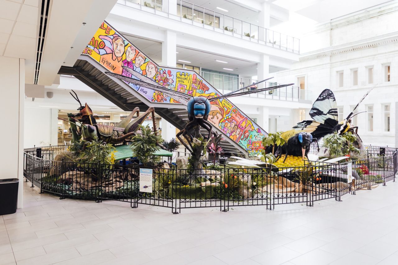 Large colorful insect sculptures including a blue-faced fly, grasshopper, bee, and butterfly inside a bright, white library atrium near a staircase with a colorful mural.