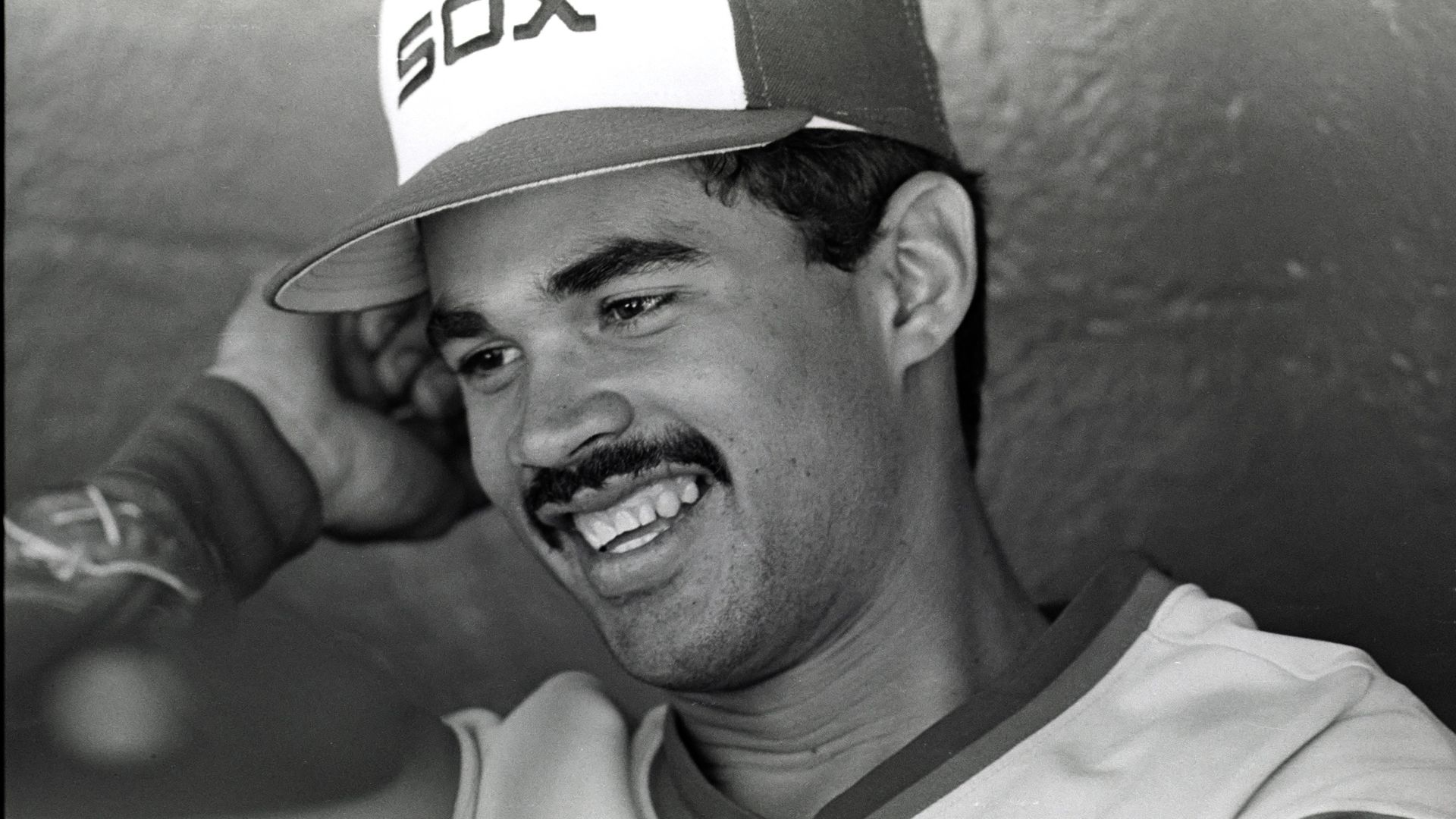 Black-and-white close-up of a smiling man wearing a baseball cap with SOX on the front and a striped jersey; background is out of focus.