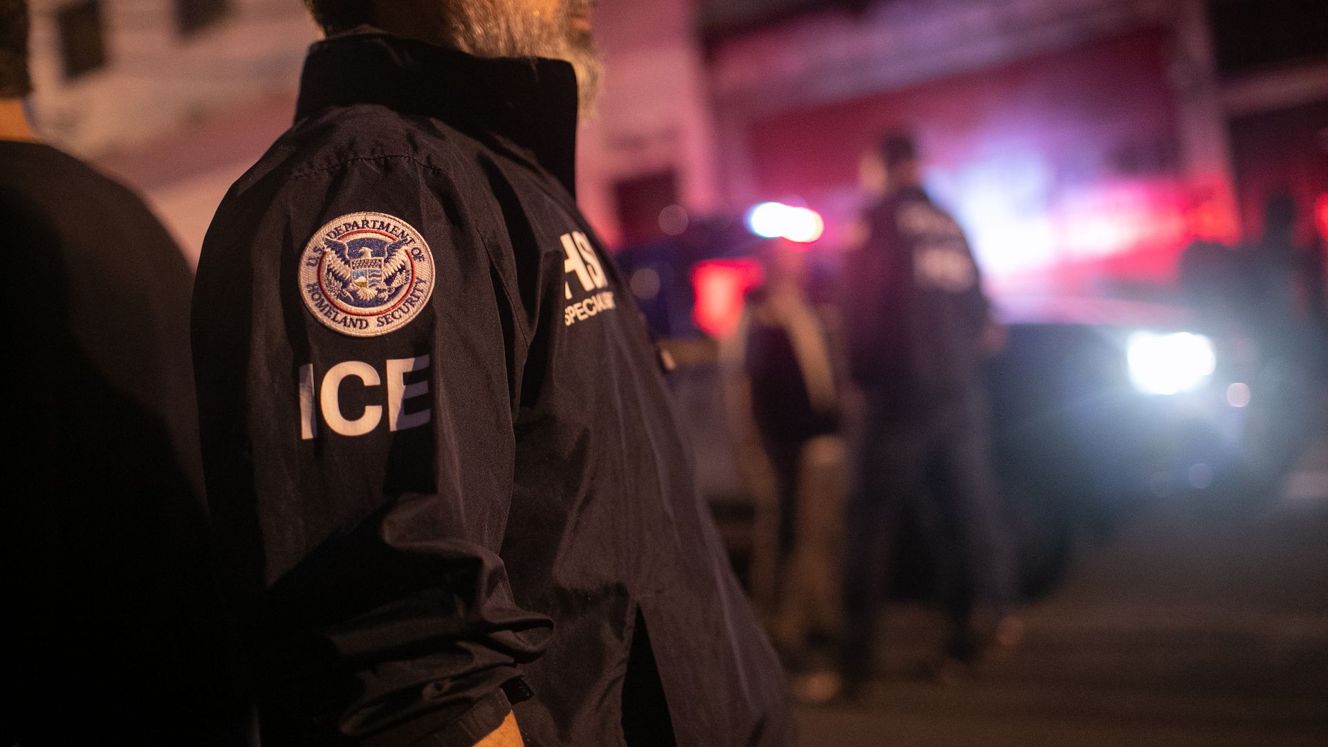 The shoulder of an ICE agent showing his patches. There are red lights in the background. 