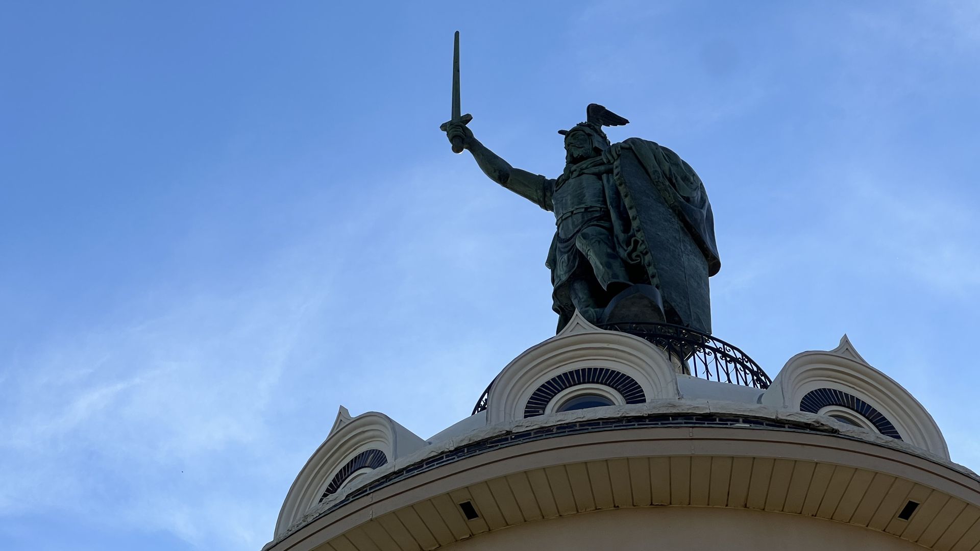 Bronze statue of a warrior with a sword raised, wearing a winged helmet and cape, standing atop a round, white building against a blue sky.