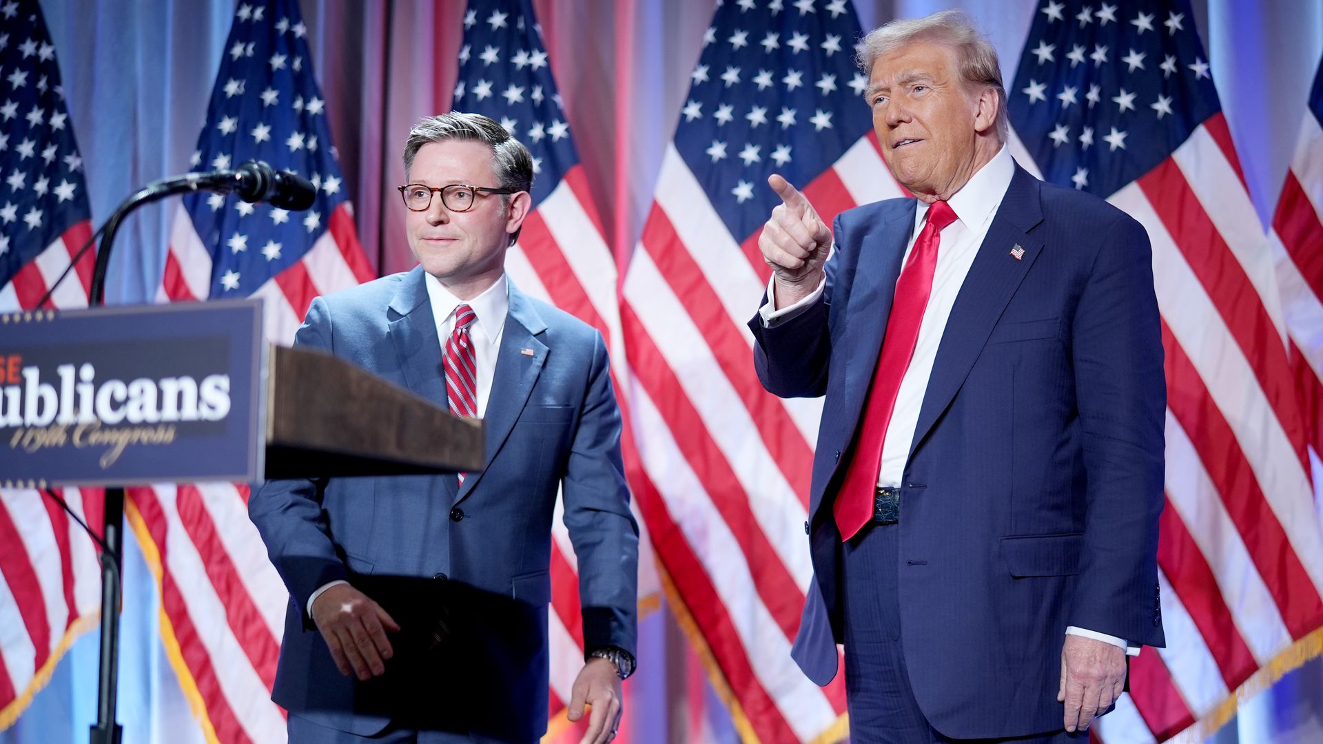House Speaker Mike Johnson and President-elect Trump, standing in front of a blue curtain and a row of American flags.
