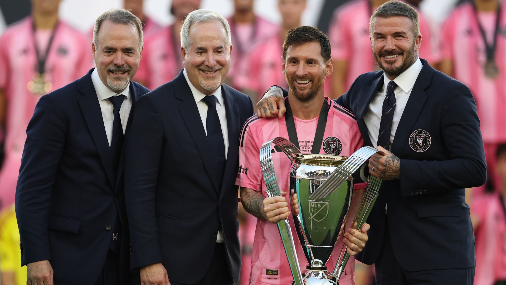 Four men pose for a photo: Lionel Messi in a pink Inter Miami jersey holding the MLS Cup, next to Jorge Mas and David Beckham in suits, with pink-clad players blurred in the background. Jose Mas is on the far left next to Jorge Mas.