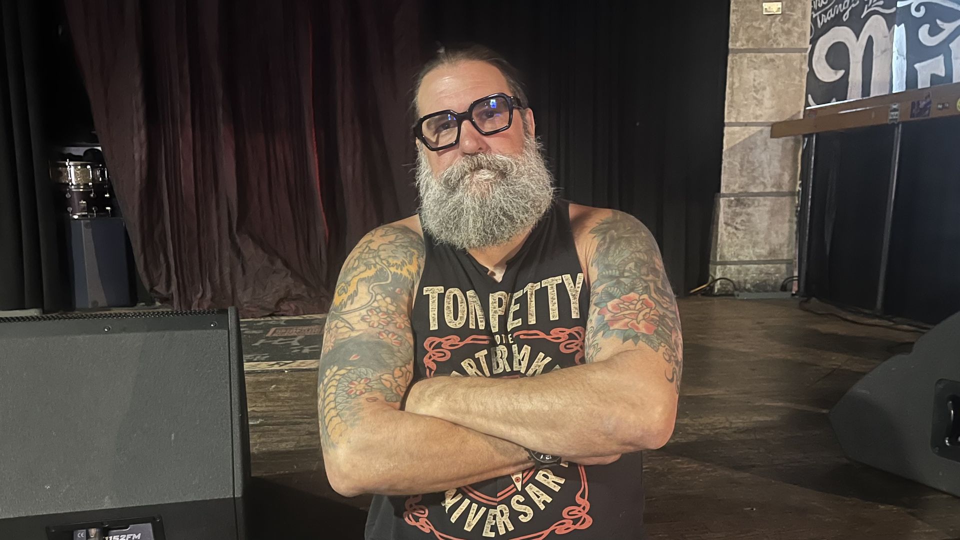 Bearded man with thick gray beard and large black glasses stands with arms crossed on a dim stage, wearing a black sleeveless shirt; tattooed arms visible in backstage setting with curtains.