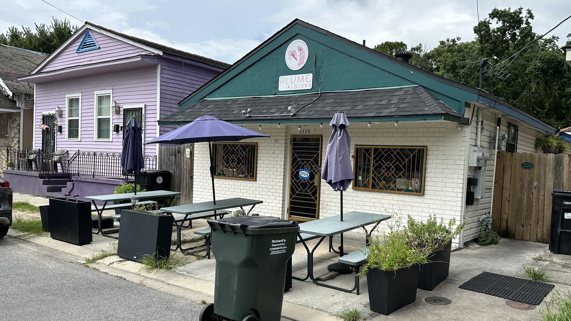 Street view of a small restaurant with green roof and white brick walls, outdoor green tables with purple umbrellas, large green trash bin, and a purple house beside it.
