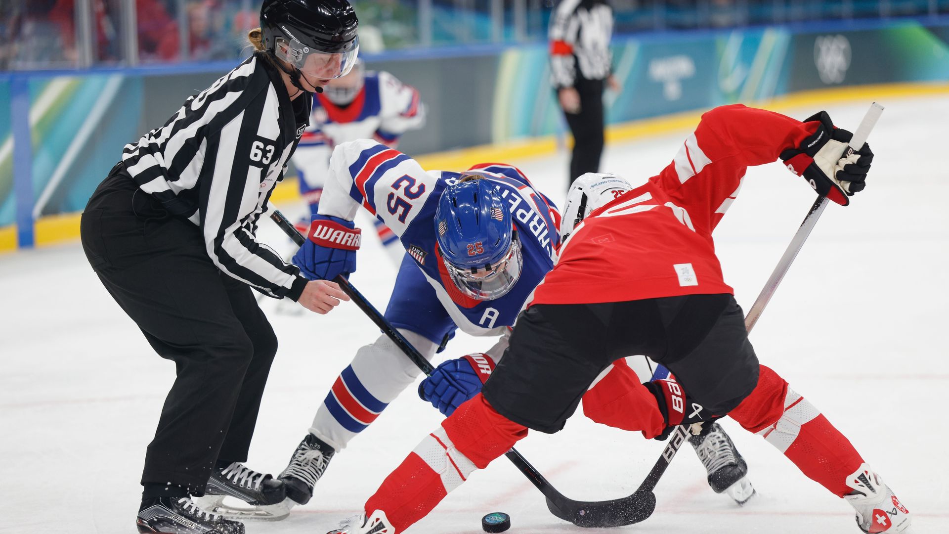 Ice hockey faceoff between a player in blue, white, and red and a player in red and black while a referee in black and white stripes drops the puck on the ice.