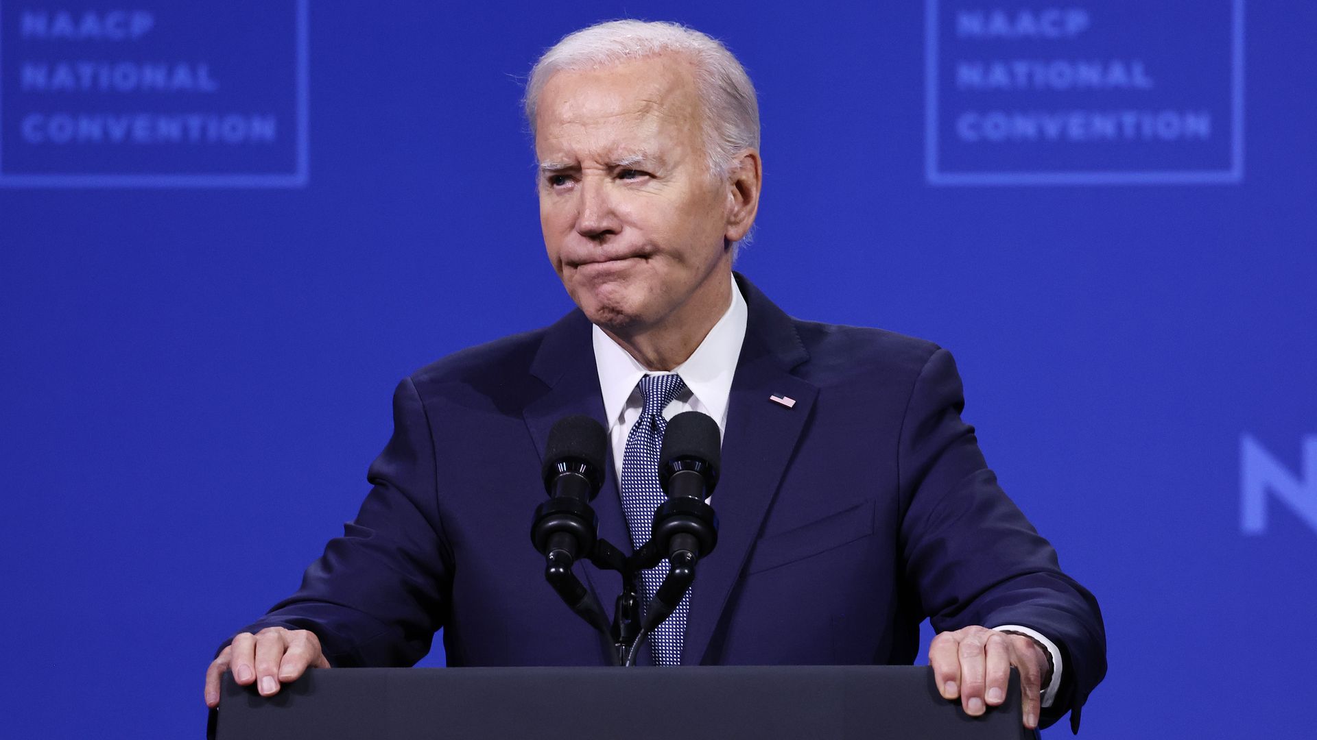 President Biden, wearing a blue suit and gripping a podium, speaking in front of a blue NAACP backdrop.