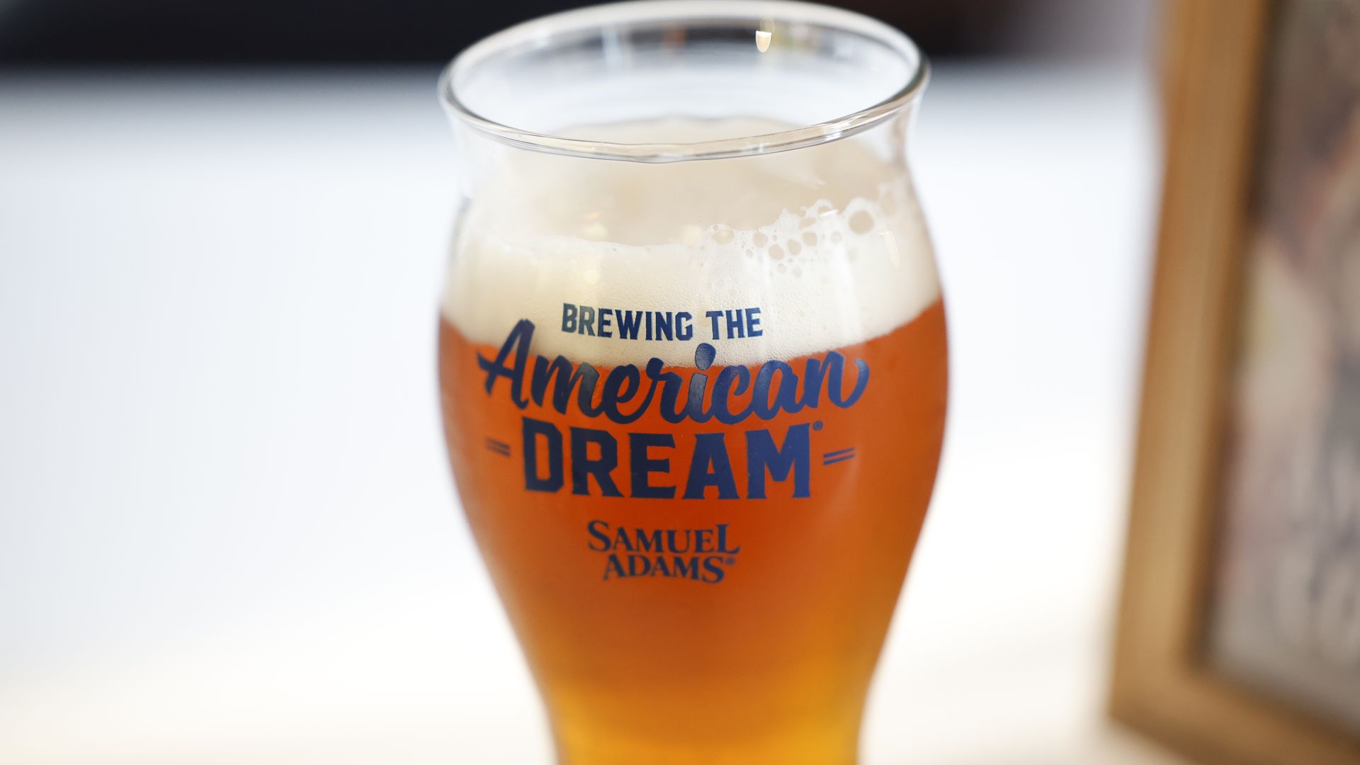 Close-up of an amber beer in a curved glass with a foamy head. Blue text on the glass reads "American Dream" with "Samuel Adams" logo below; blurred background.
