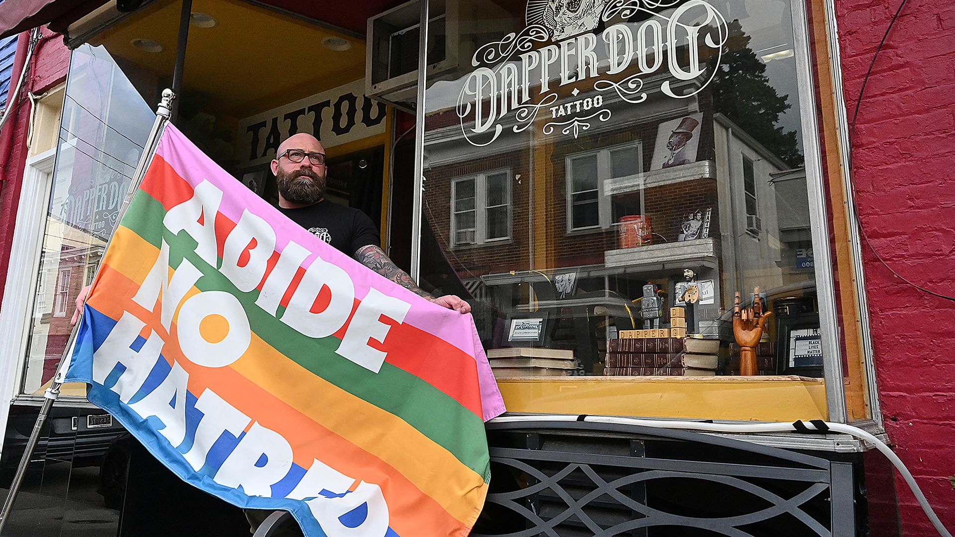 A bearded man with tattoos holds a rainbow-striped flag that says "ABIDE NO HATRED" outside Dapper Dog tattoo shop with a large window and red brick wall.