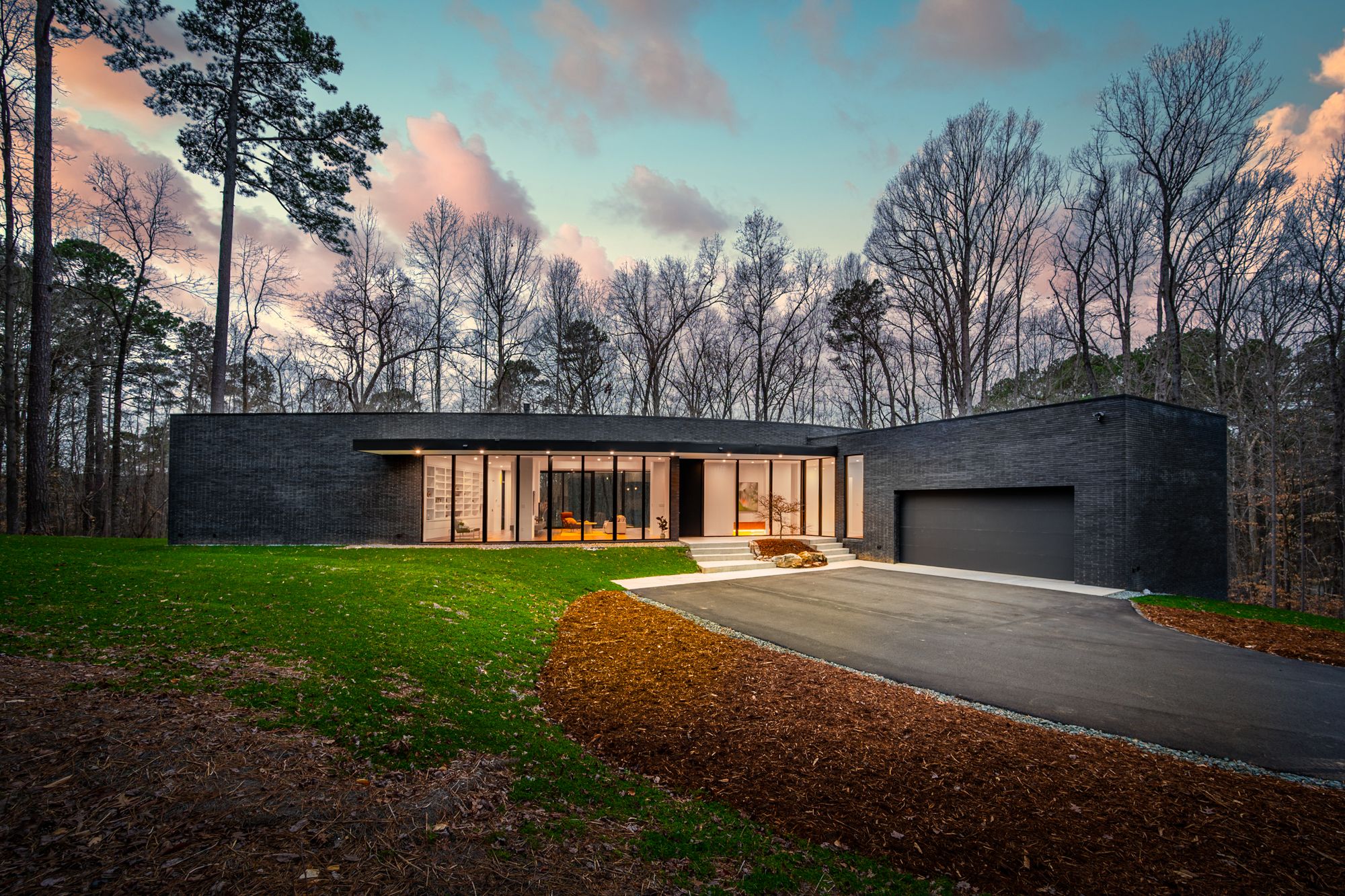 Modern black brick house with large glass windows and garage, set against bare trees and a colorful sky at dusk, with green grass and brown mulch landscaping.