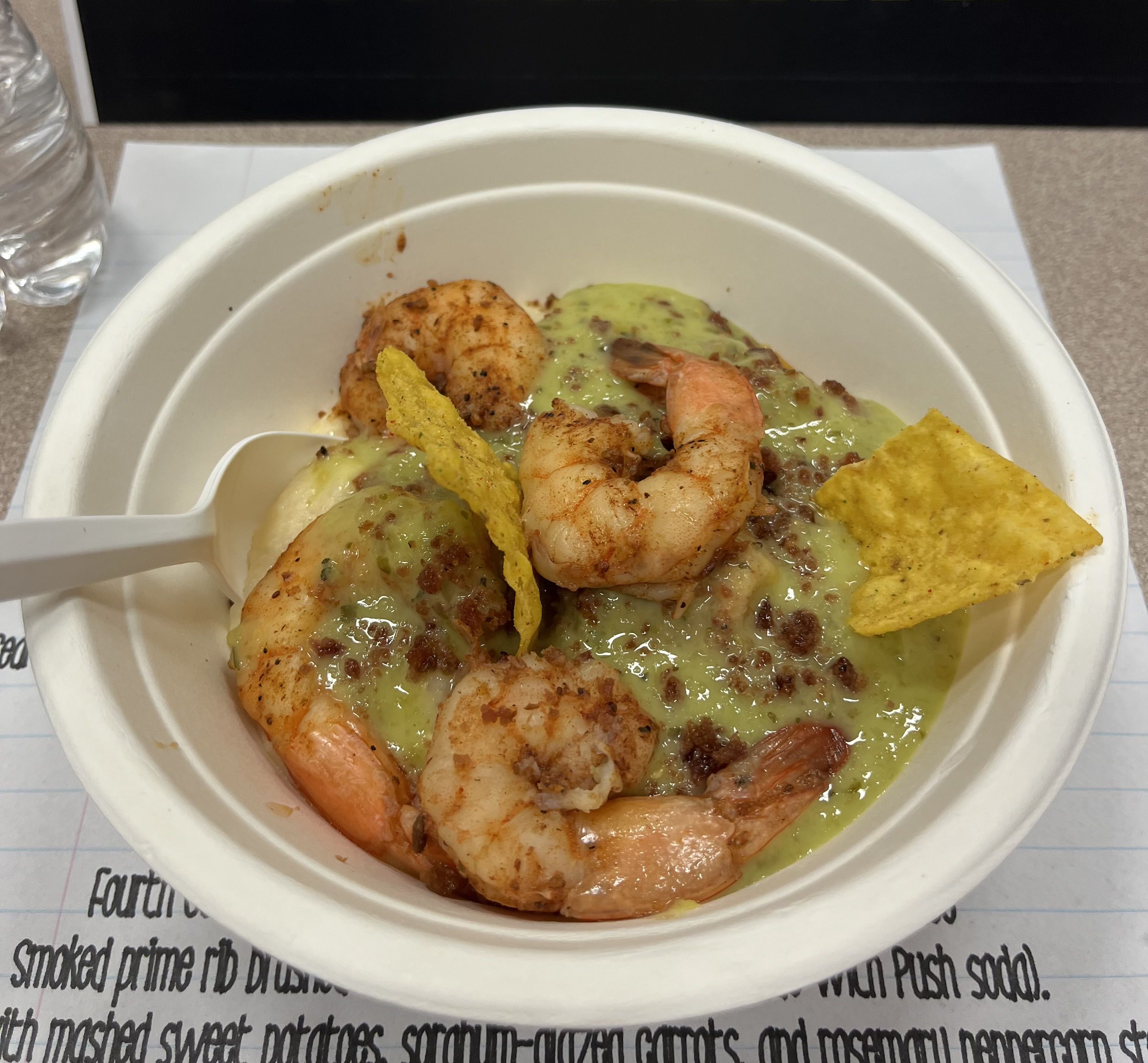 Bowl with cooked shrimp, creamy green sauce, crumbled bits, and two yellow tortilla chips, with a plastic spoon on the side.