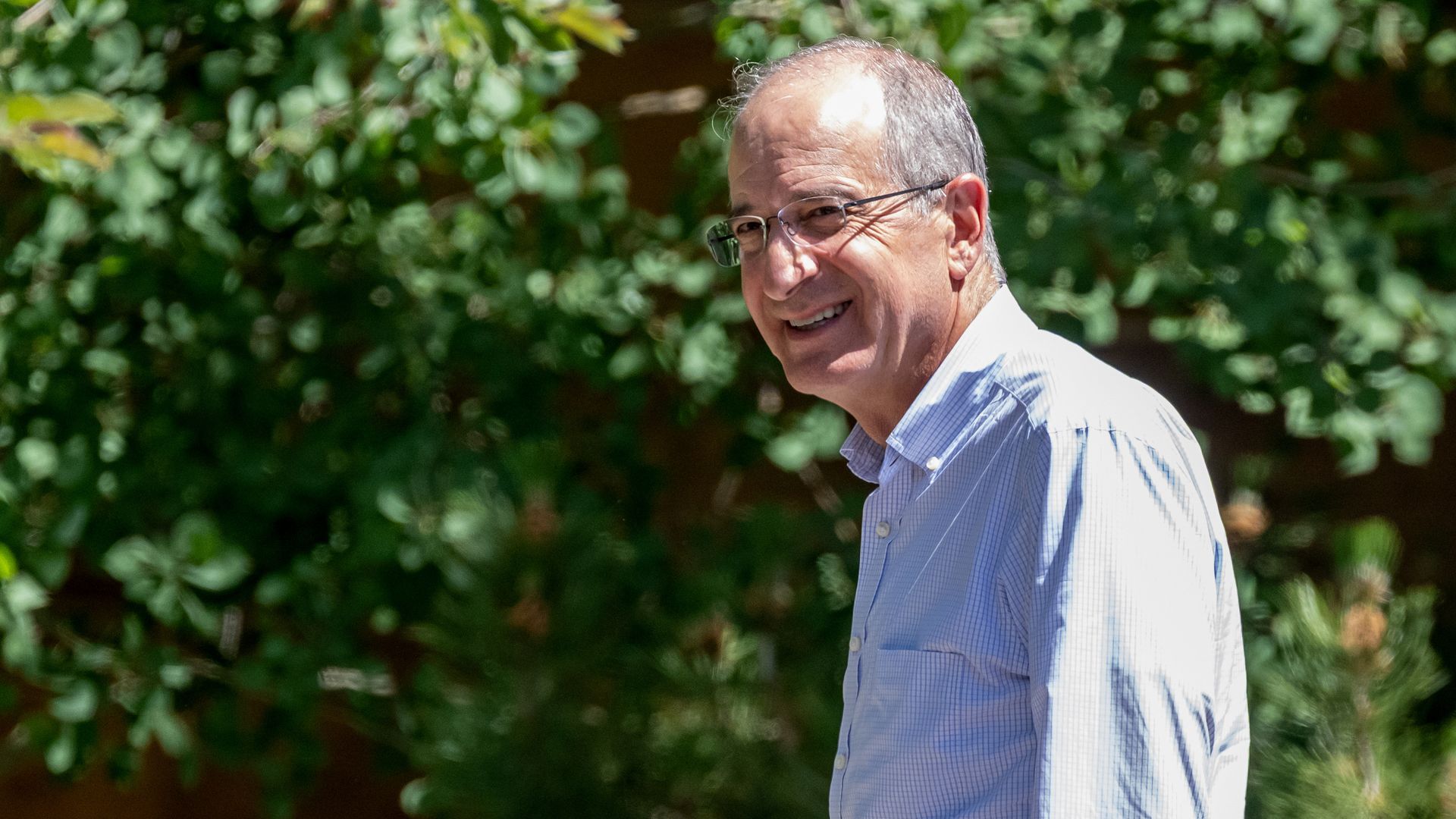 A person in a white collared shirt and glasses walks outside near some bushes