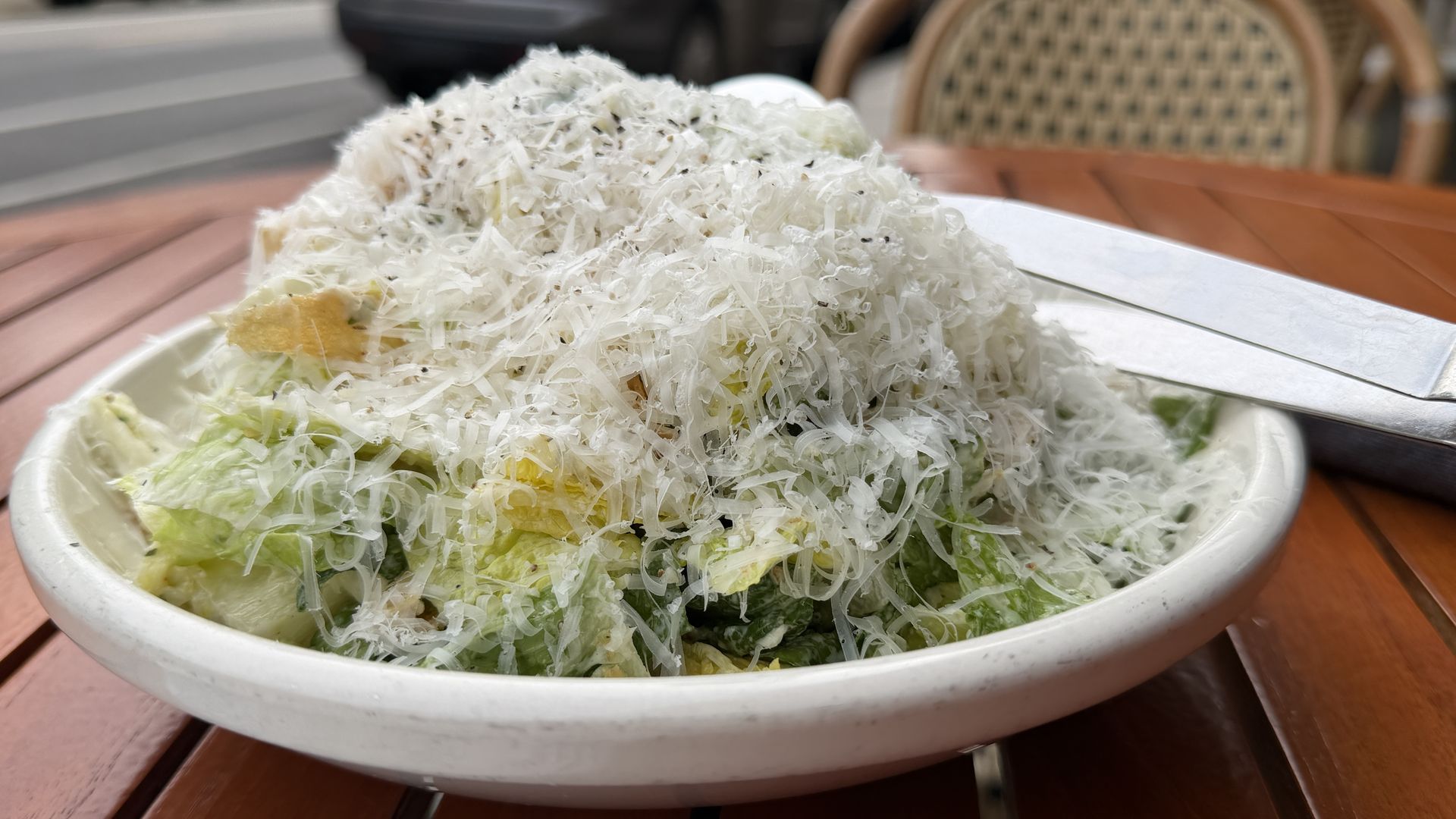 Large mound of shredded white cabbage salad piled in a white bowl on a wooden outdoor table, with egg pieces; blurred cars and a woven chair in the background.