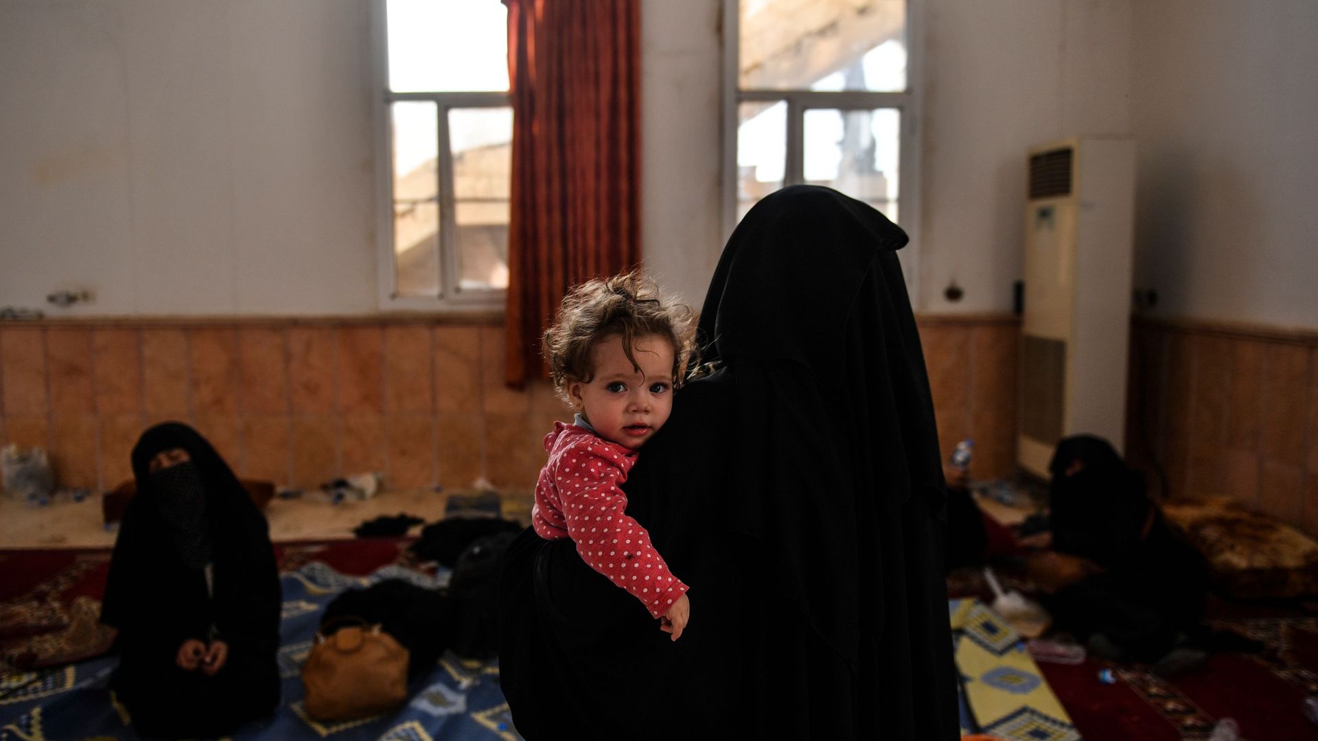 A woman holds a child in an open room.