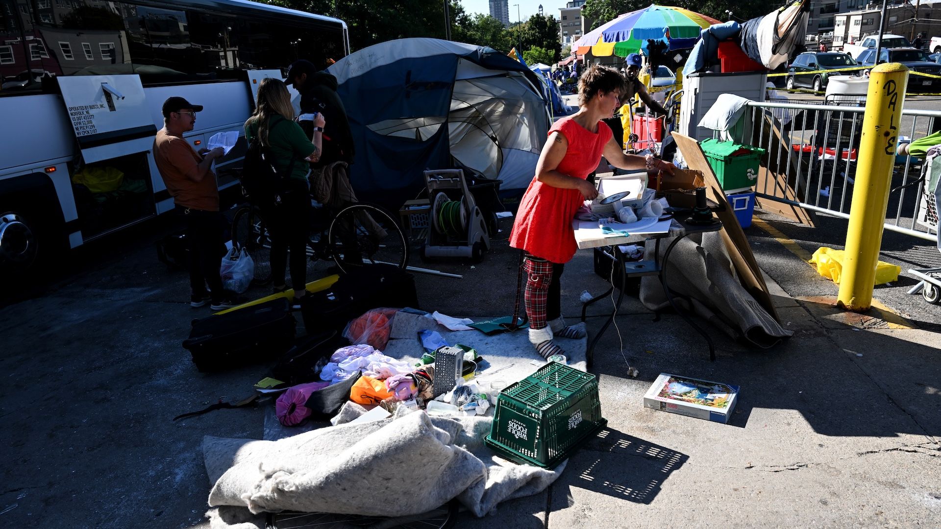 Sunlit street camp with tents and a colorful umbrella vendor booth. A woman in a red dress works at a cluttered table of papers and items while bystanders browse near a bus and yellow pole.