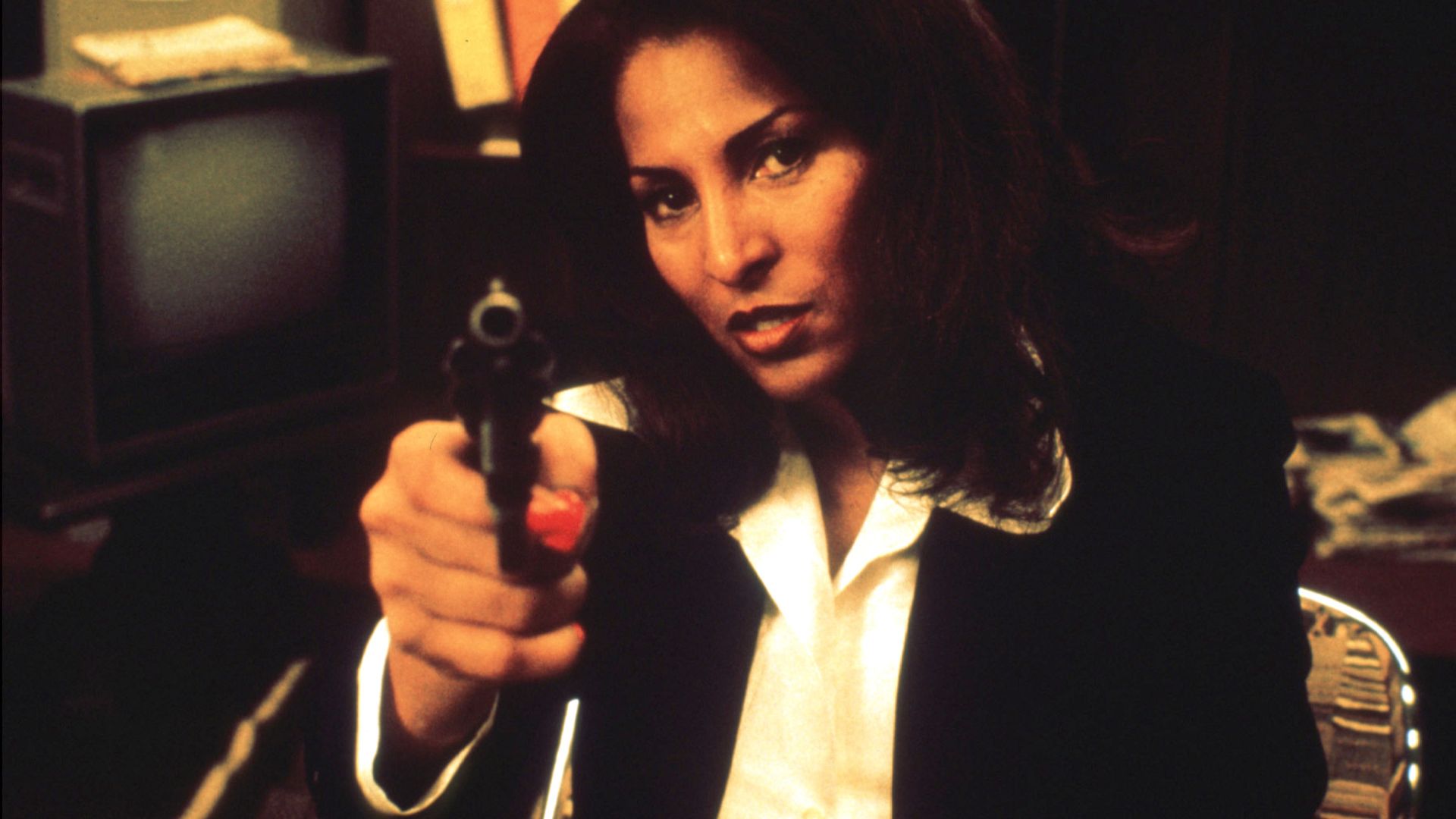 A woman with dark hair, wearing a white blouse and black blazer, points a handgun toward the camera in a dim room; a vintage TV and shelves are visible in the background.