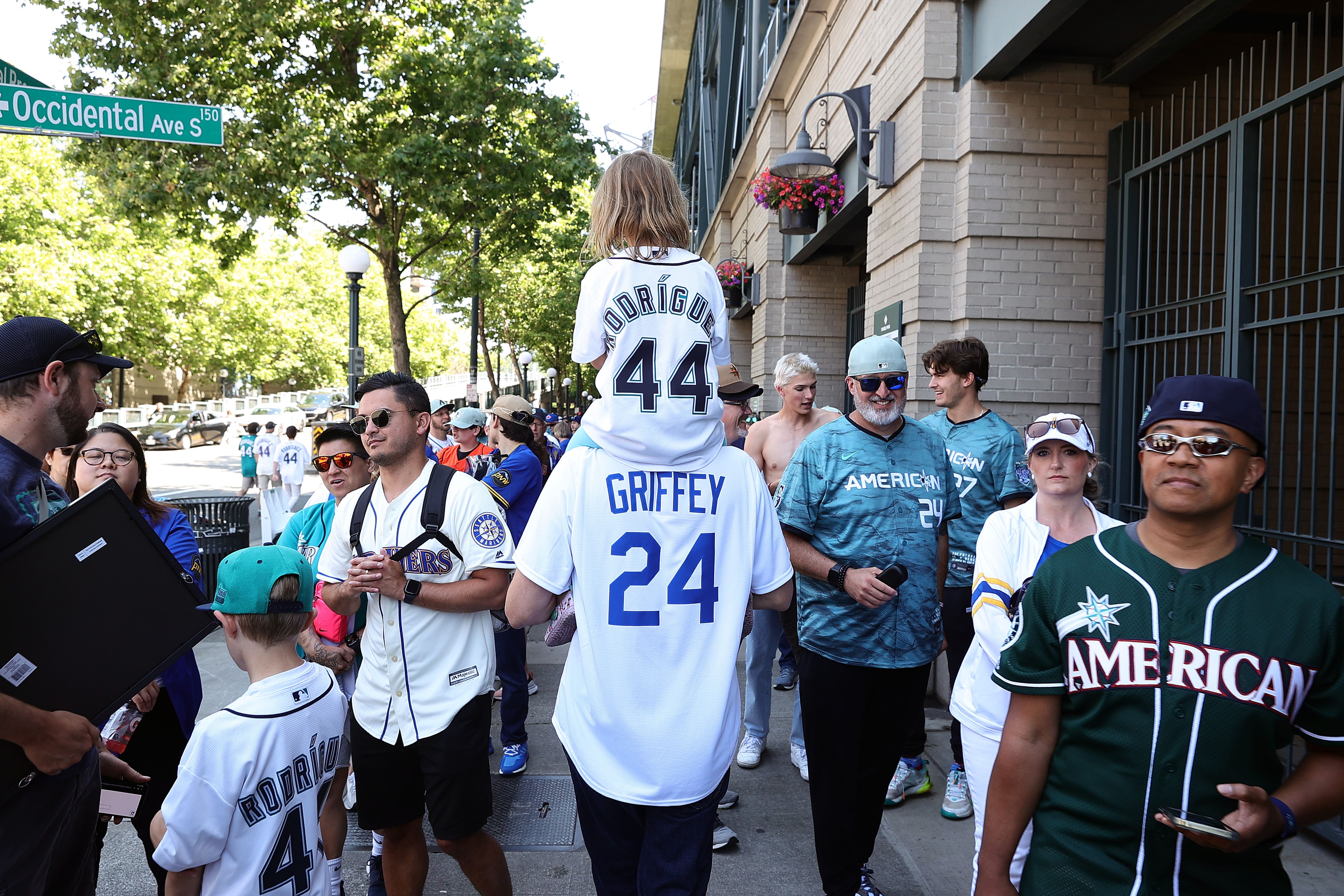 A man wearing a Ken Griffey jersey holds a girl on his shoulders who is wearing a Julio Rodriguez jersey as other fans walk by them on the sidewalk.