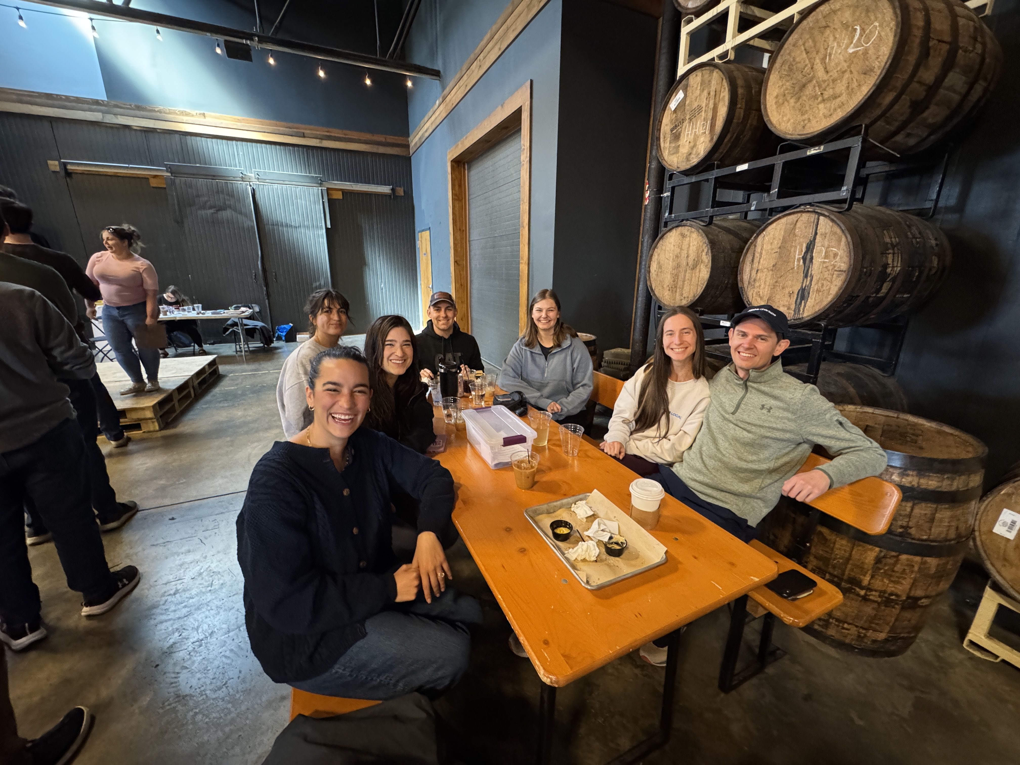 Seven people sitting at a wooden table in a brewery-like setting with barrels on shelves behind them, smiling and enjoying drinks with casual clothing indoors.