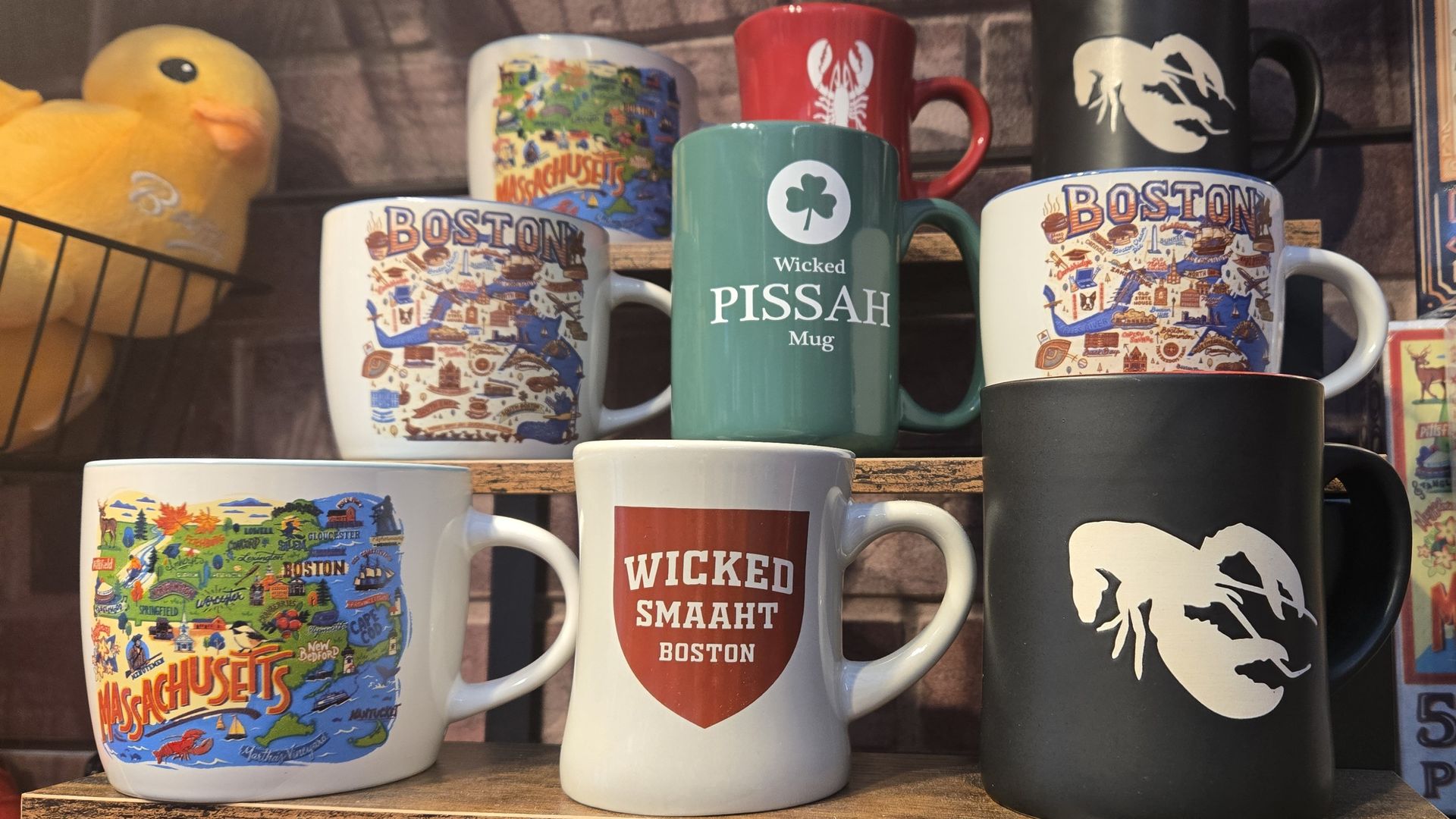 A display of souvenir mugs featuring Massachusetts and Boston themes, including colorful maps, lobsters on red and black mugs, and mugs with phrases "Wicked Pissah" and "Wicked Smaaht Boston."