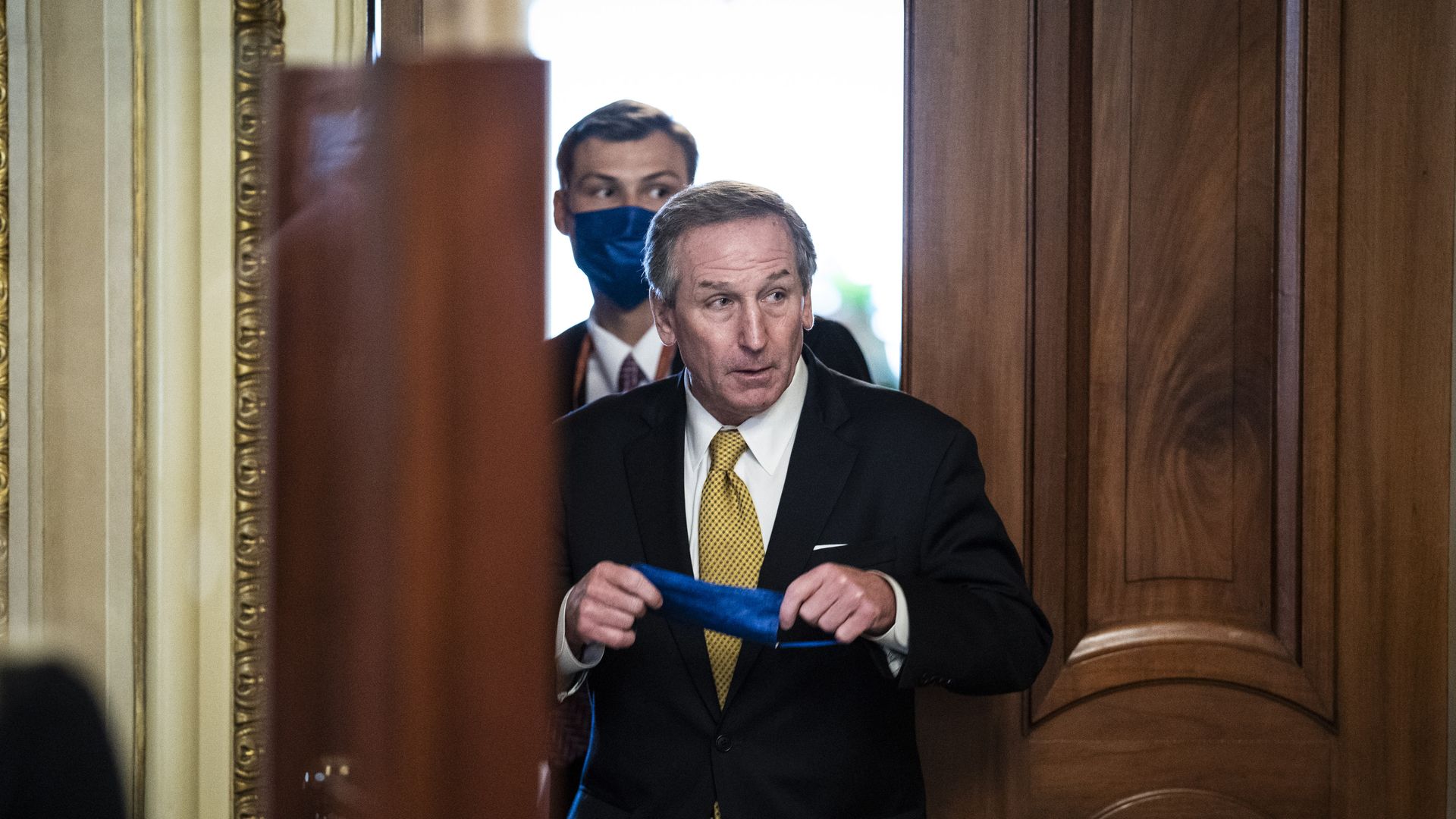 Lawyer Michael van der Veen walks through a Senate doorway with his mask in his hands and a masked man behind him.