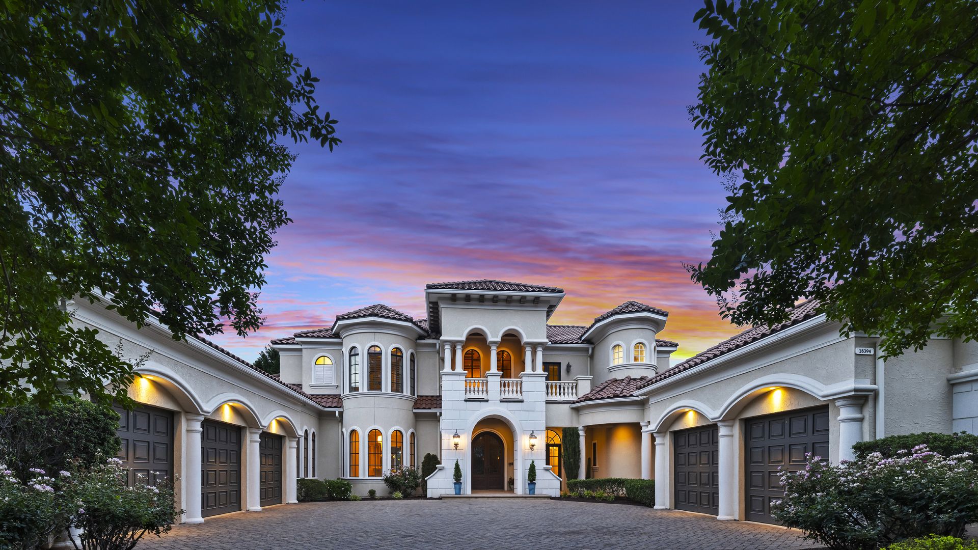 Mediterranean-style mansion with arched windows and tiled roofs, a cobblestone courtyard, and lush trees framing the scene as a blue, pink, and orange sunset colors the sky.
