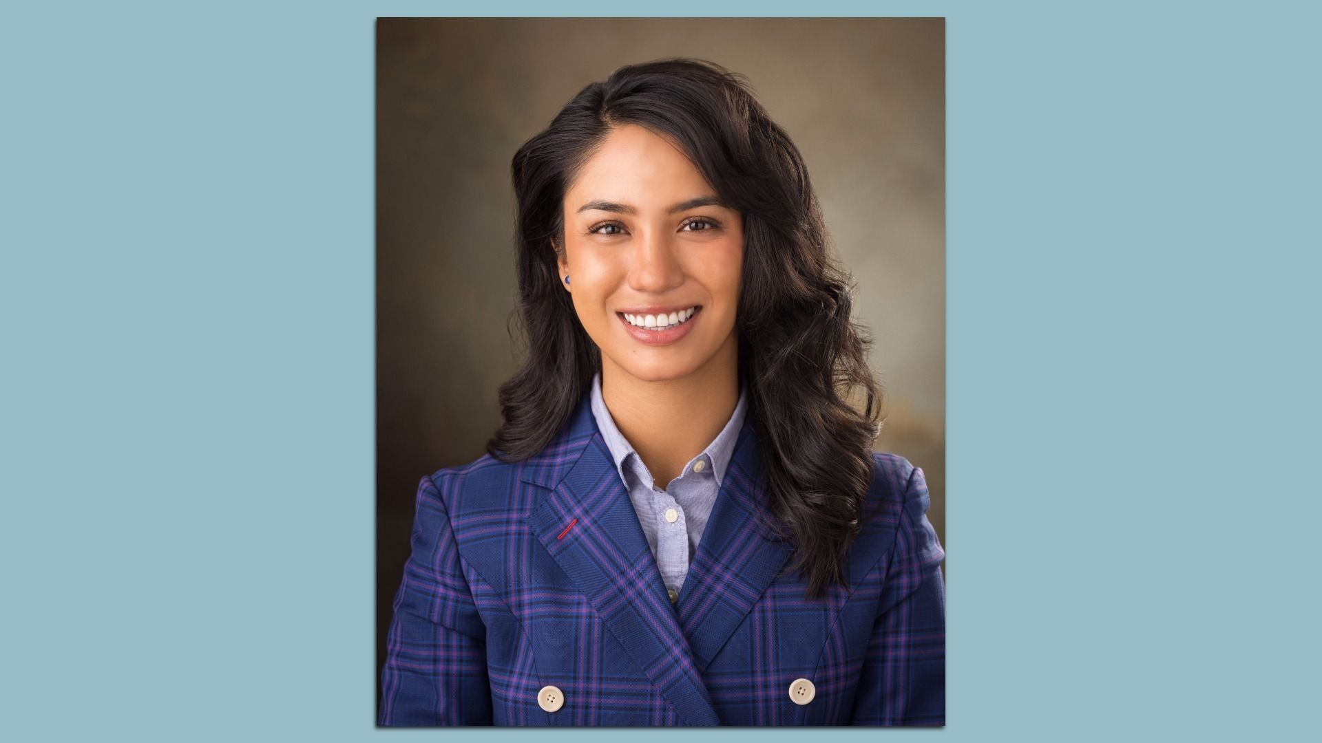 Portrait of a smiling woman with dark, wavy hair wearing a blue plaid blazer over a light blue shirt, posed against a neutral brown gradient backdrop.