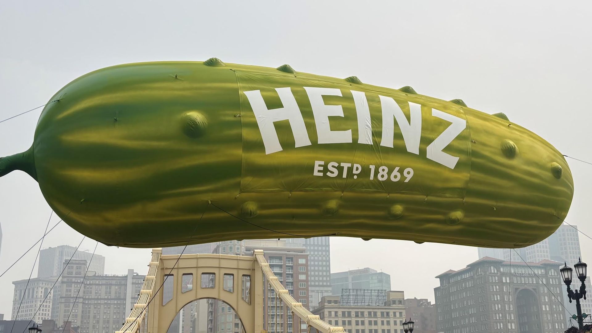Green blimp with white "HENZ" and "EST 1869" drifts over a city skyline, above a yellow arch bridge; cables and tall buildings are visible in the background.