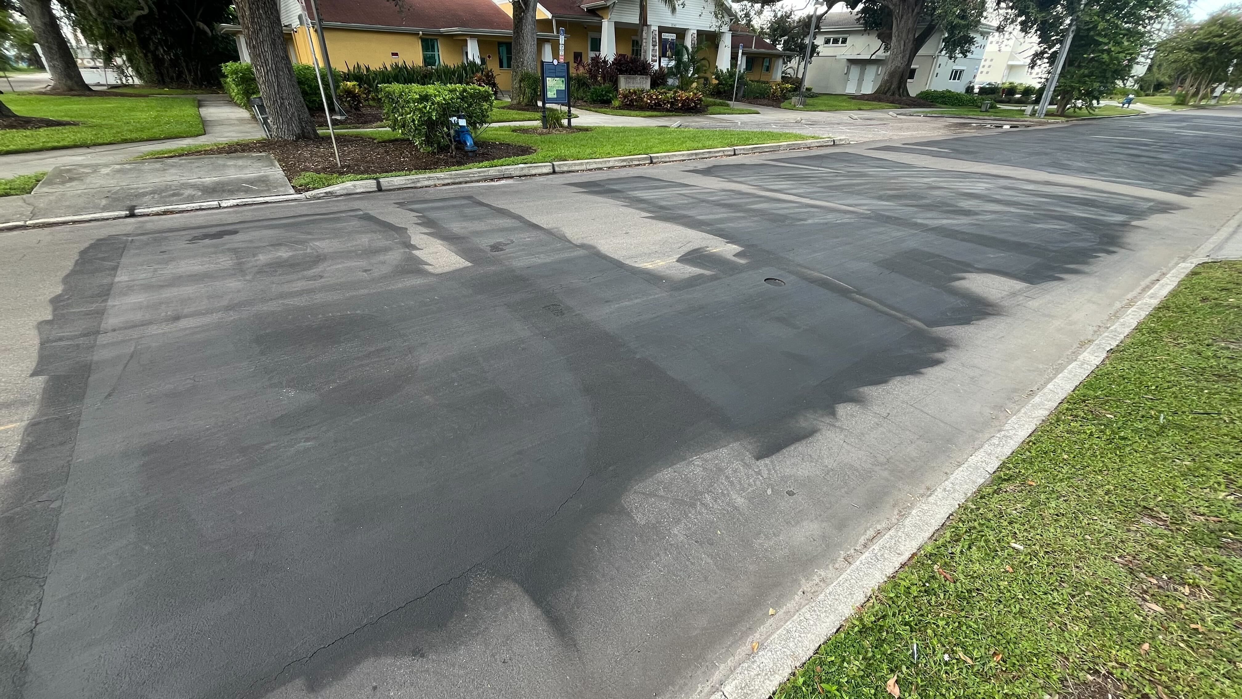 A residential street with freshly patched black asphalt covering multiple rectangular sections. Green grass and sidewalks flank the road, with houses and trees in the background.