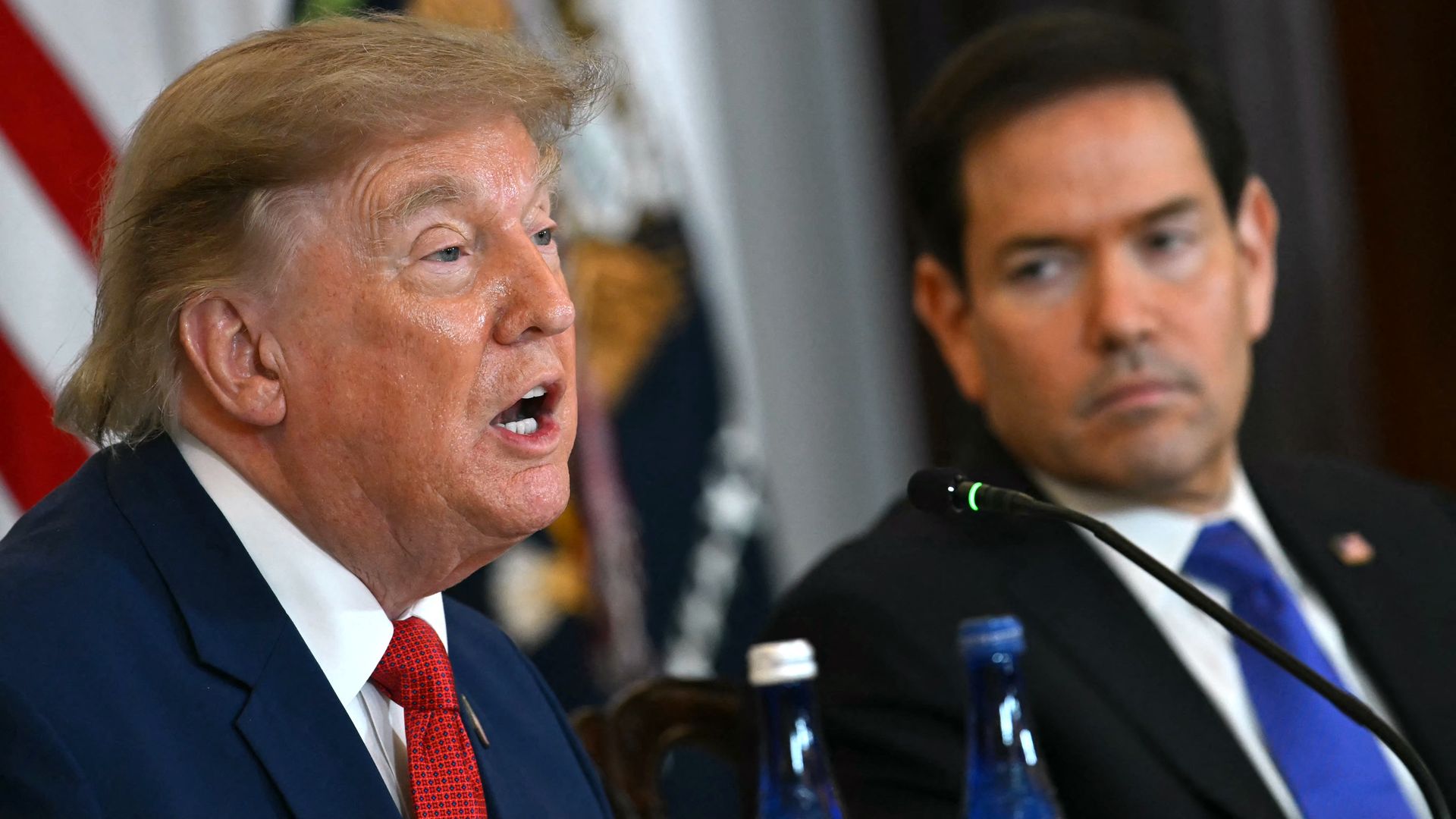 Close-up of former U.S. President Donald Trump speaking into a microphone, wearing a navy suit and red tie, with a man beside him and blurred flags in the background.