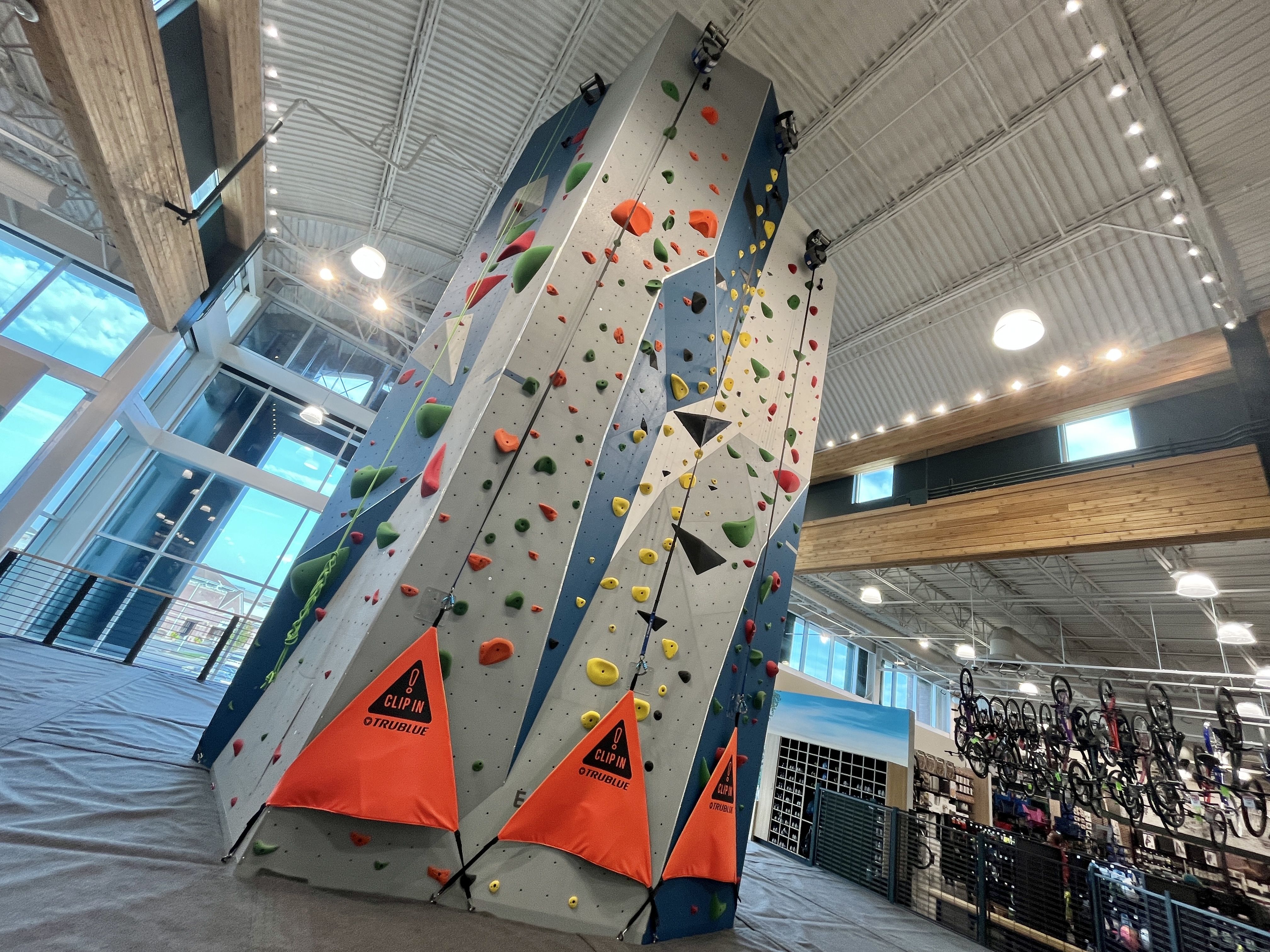 Indoor rock climbing wall with colorful holds in red, green, yellow, and black inside a spacious gym with large windows and hanging bicycles on the right side.