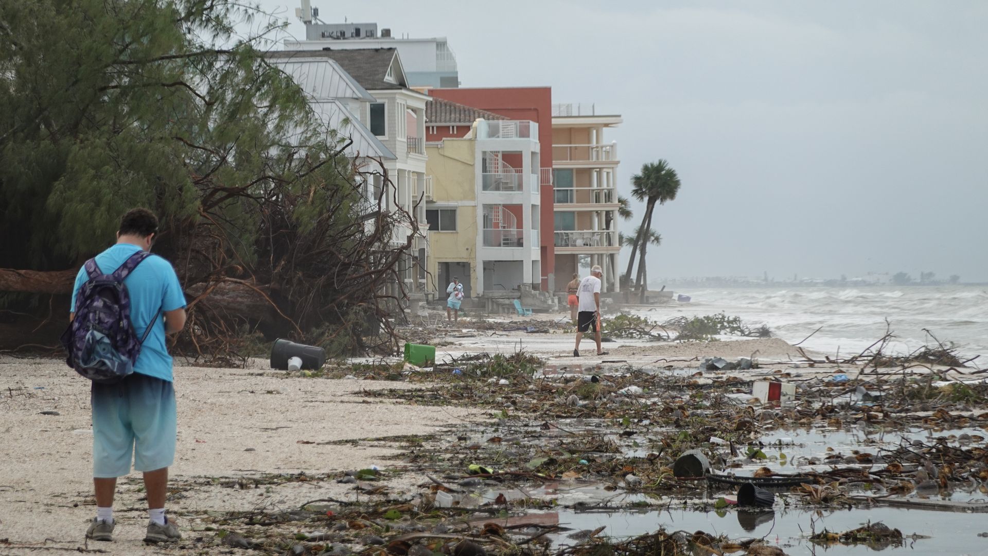 A shoreline covered in debris including tree branches, plastic plant pots and garbage bins. A man in a teal shirt with a purple backpack looks on.