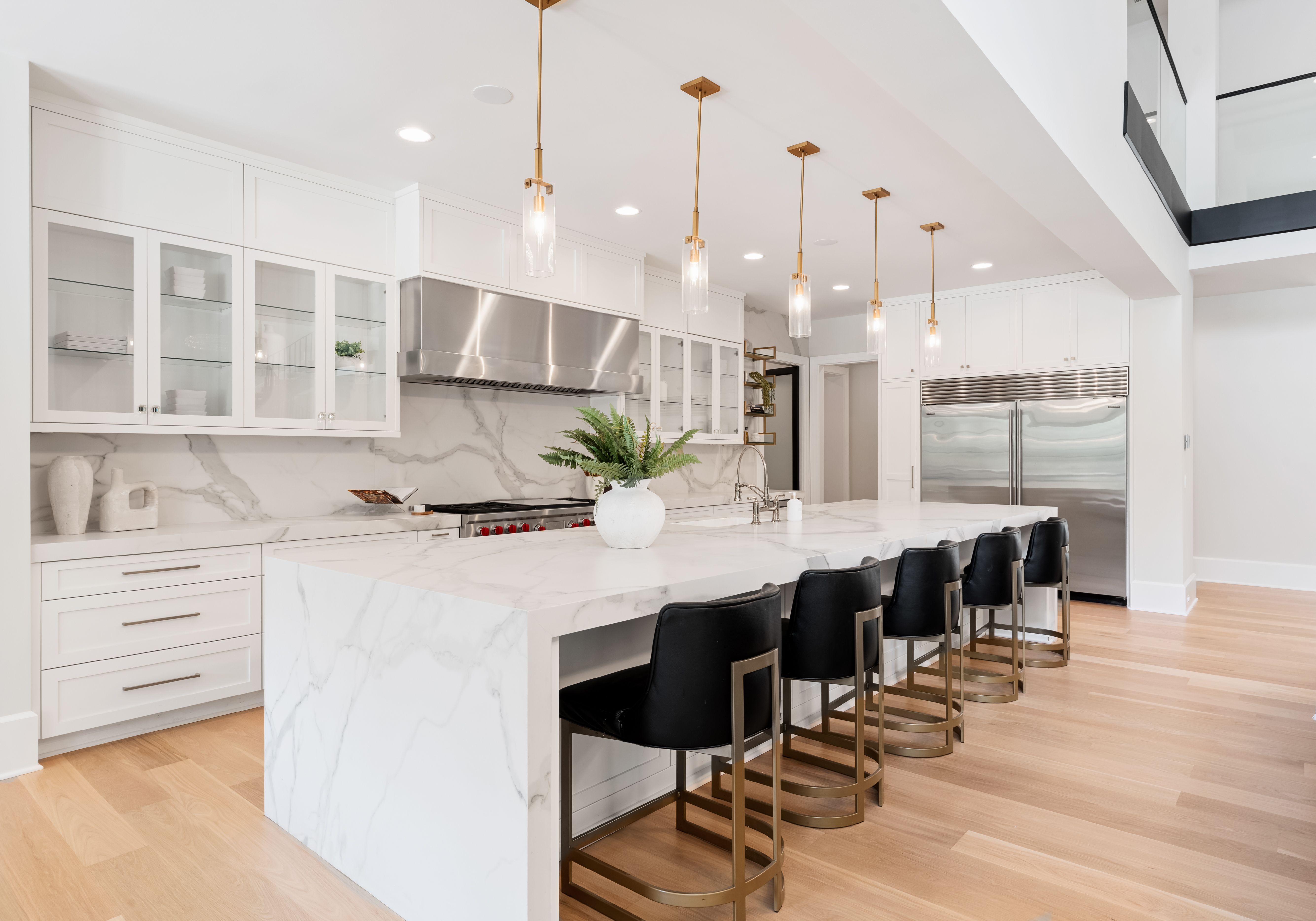 Modern kitchen with white marble island, black leather bar stools, brass pendant lights, glass cabinets, stainless steel appliances, and light wood floors.