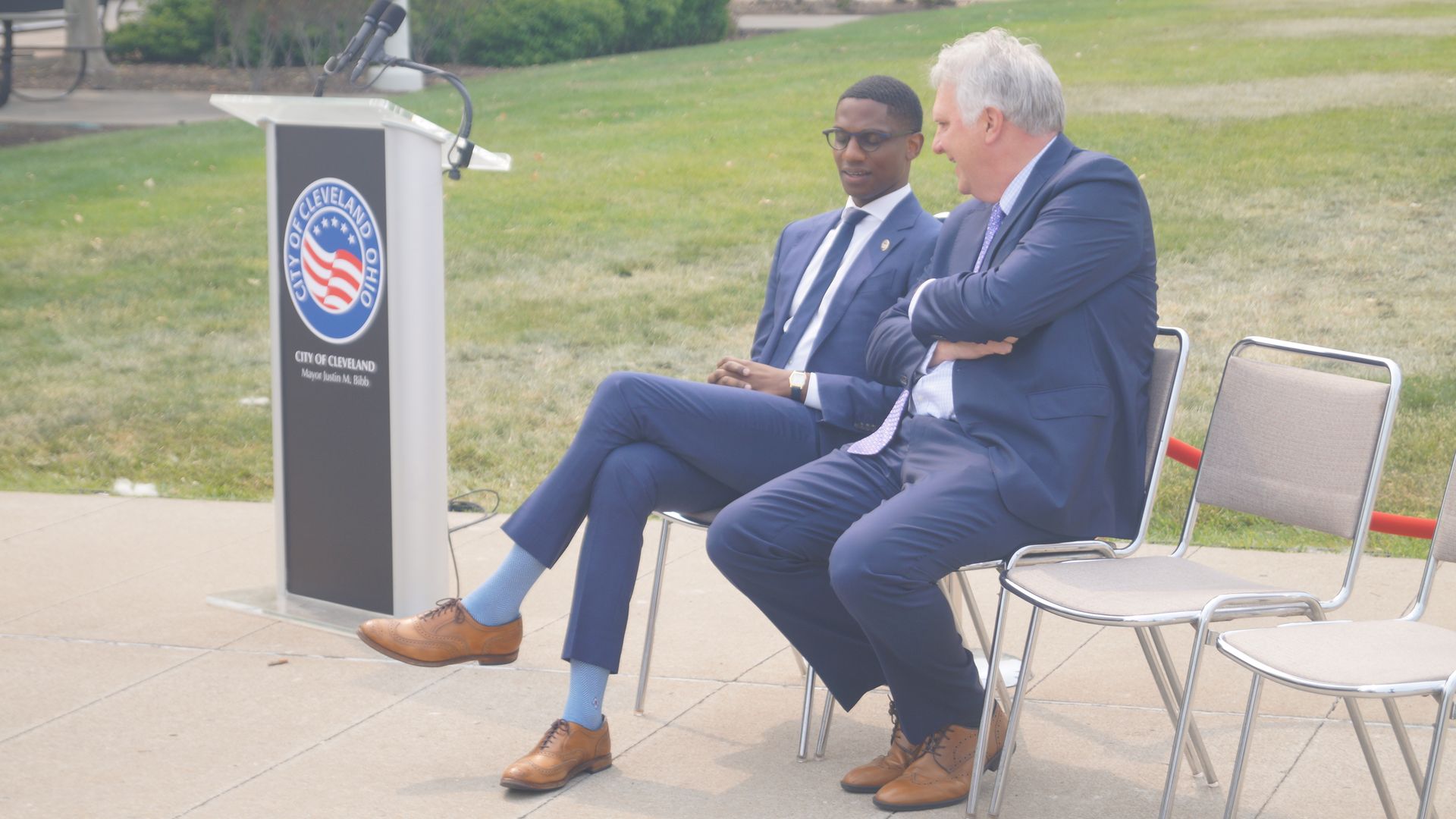 Photo of two men in suits sitting next to an outdoor podium