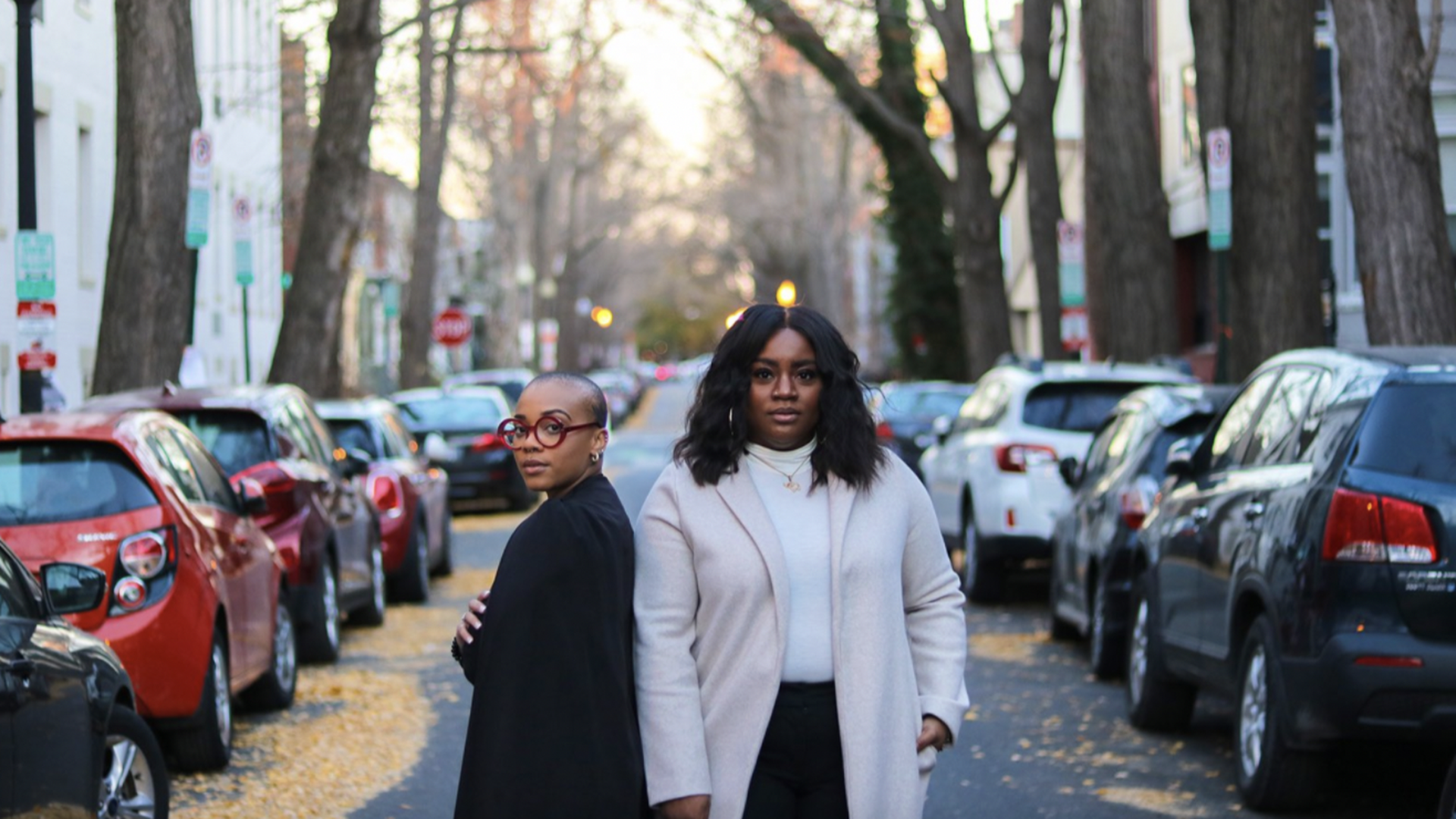 Two women stand in the middle of a tree-lined street back-to-back