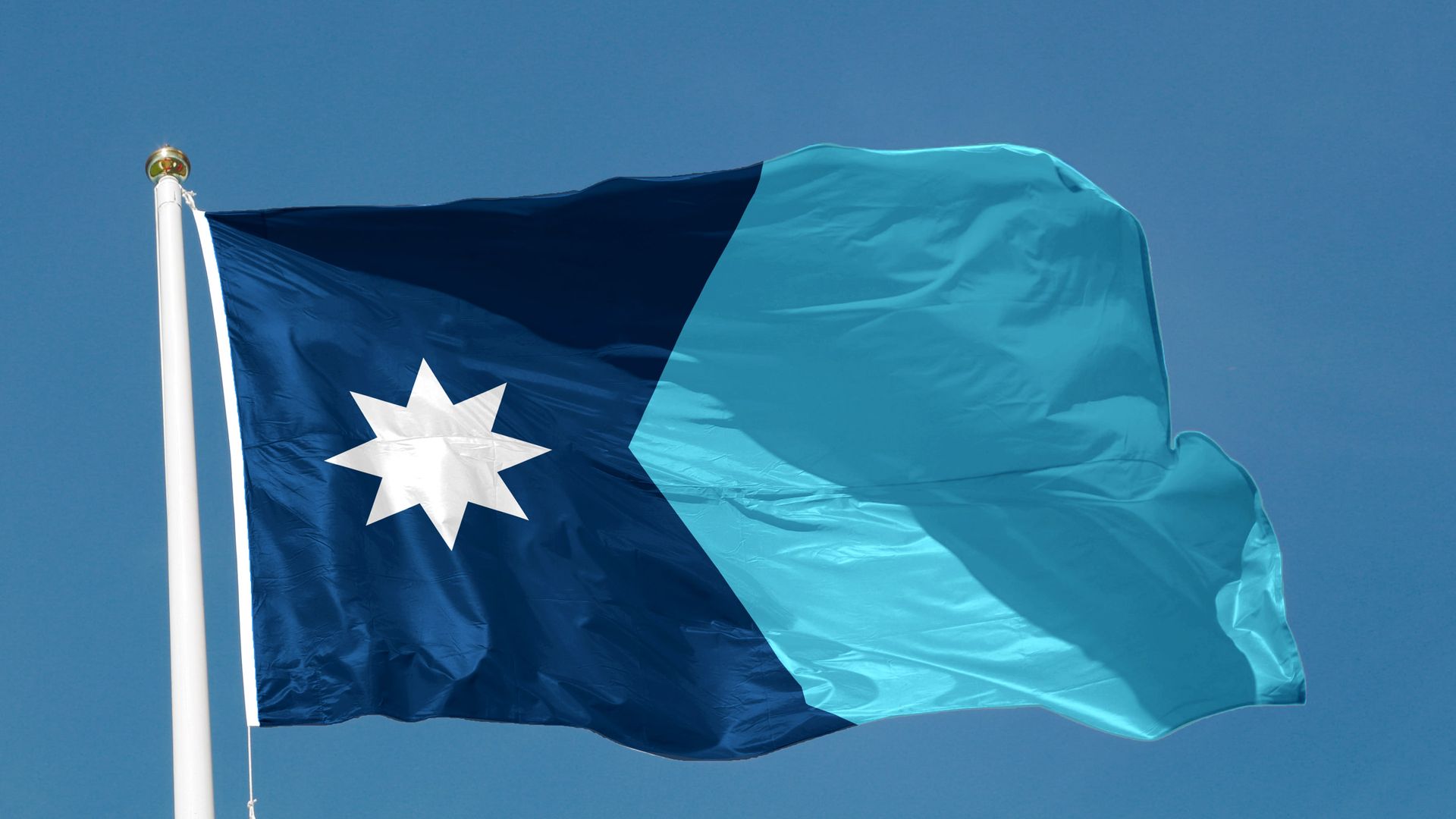 Flag waving on a white pole: dark blue left panel with a white eight-pointed star, light blue right panel; fluttering against a clear blue sky.