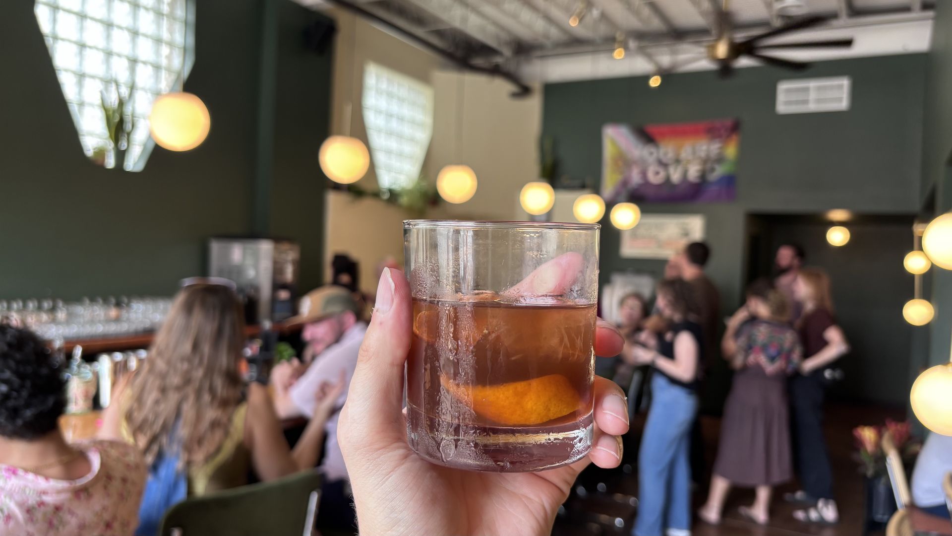 Close-up of a hand holding a glass with ice and an orange slice, in a lively bar with warm hanging bulbs and blurred patrons in the background.
