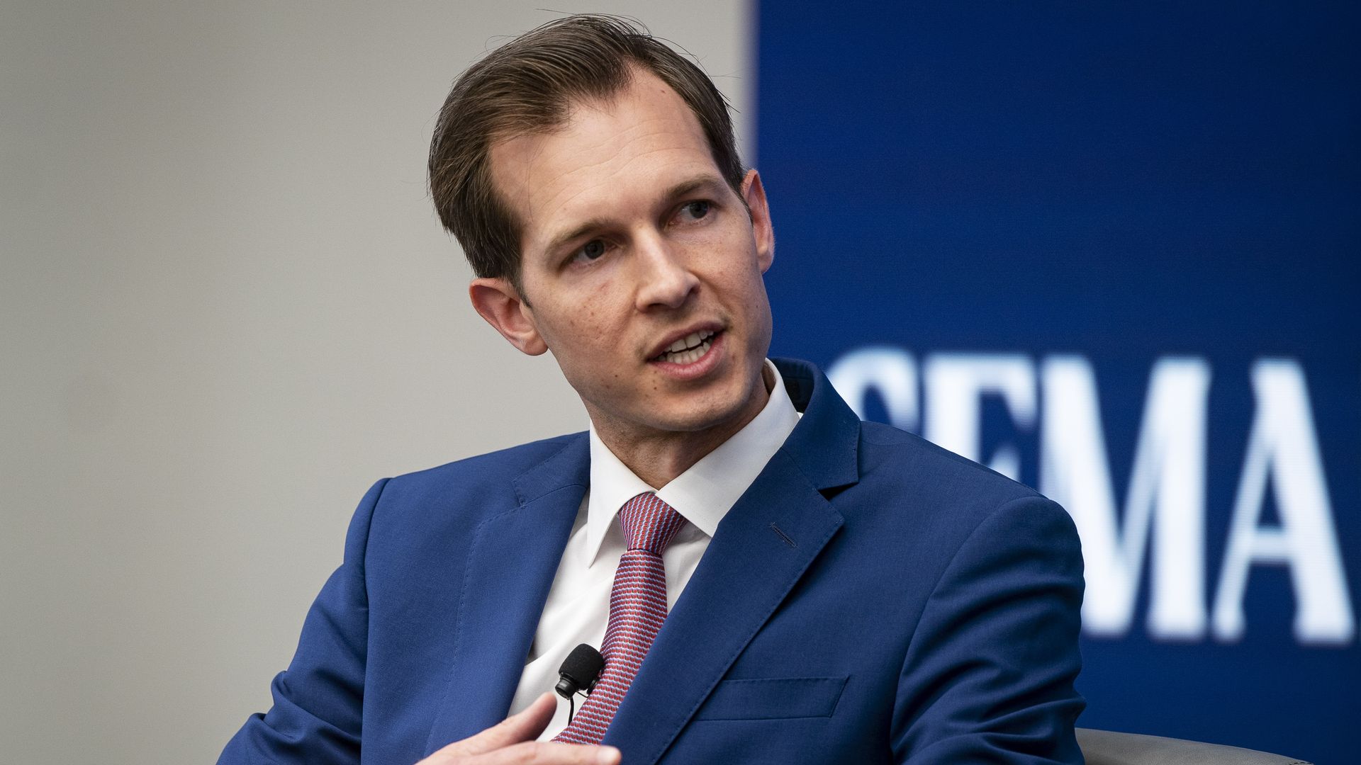 Rep. Jake Auchincloss, wearing a blue suit and sitting in a black leather chair in front of a blue backdrop.