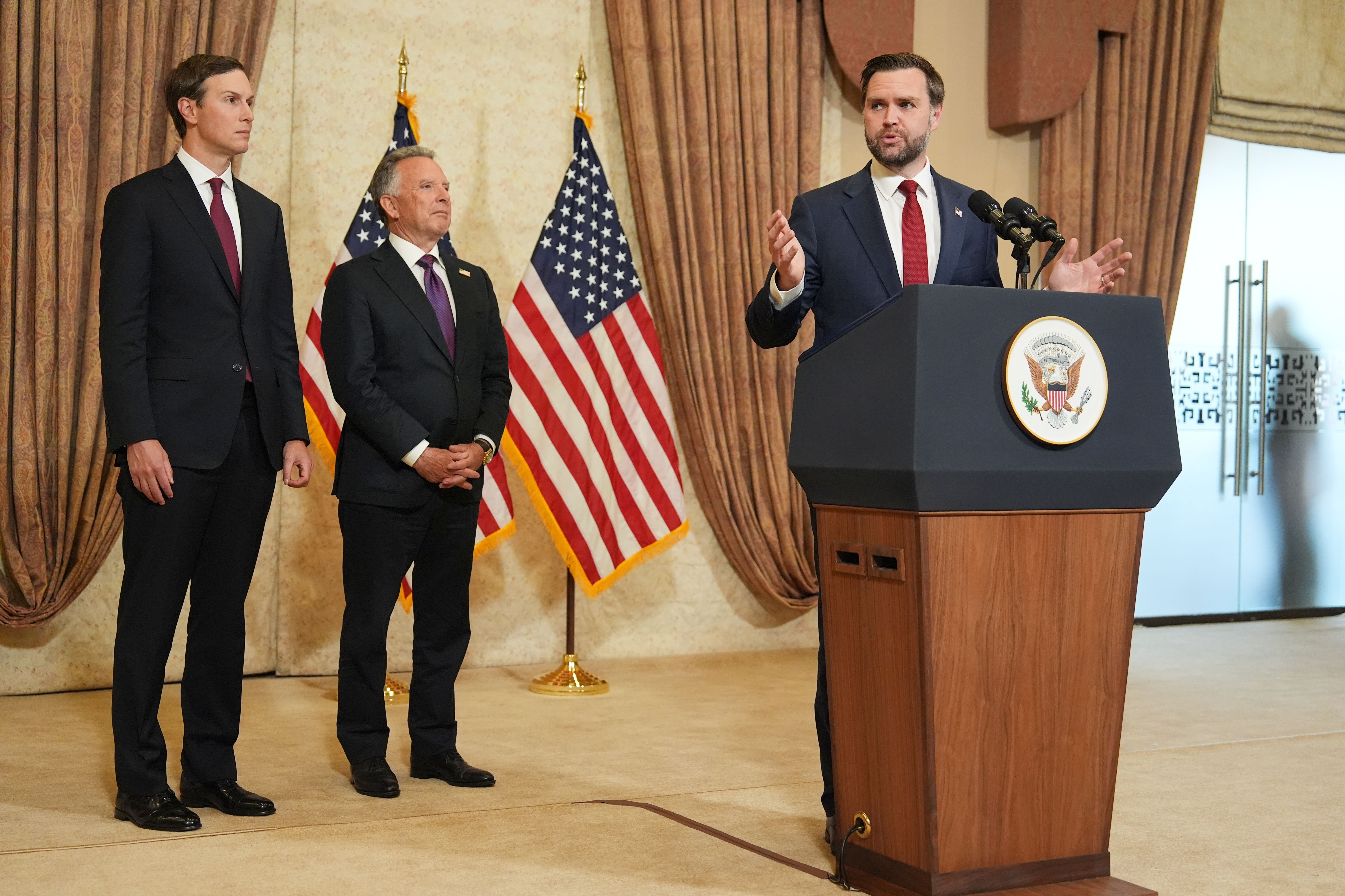 Men in suits in a news conference room with one at a podium. American flag in background