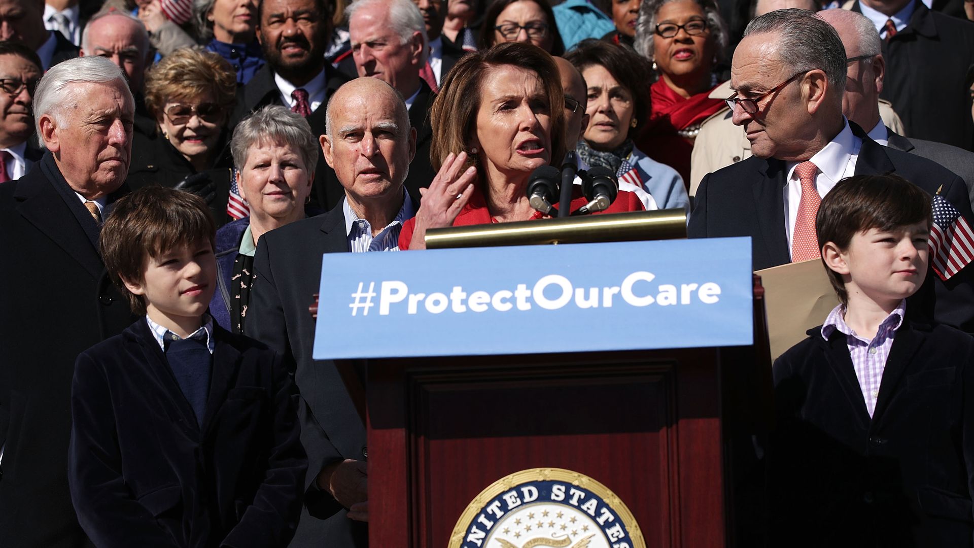 In this image, Nancy Pelosi stands outside with a crowd of lawmakers behind her, at a podium with a sign that reads "#ProtectOurCare."