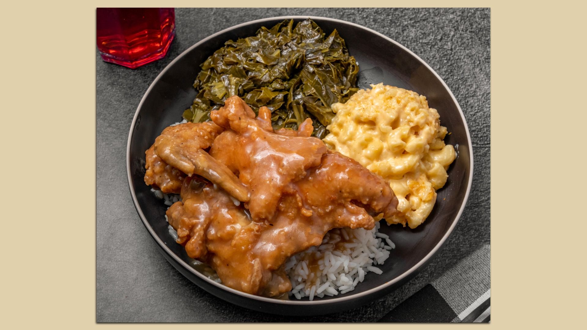 Plate with fried chicken wings in brown gravy over white rice, cooked collard greens, and creamy macaroni and cheese on a dark surface with a red drink partially visible.