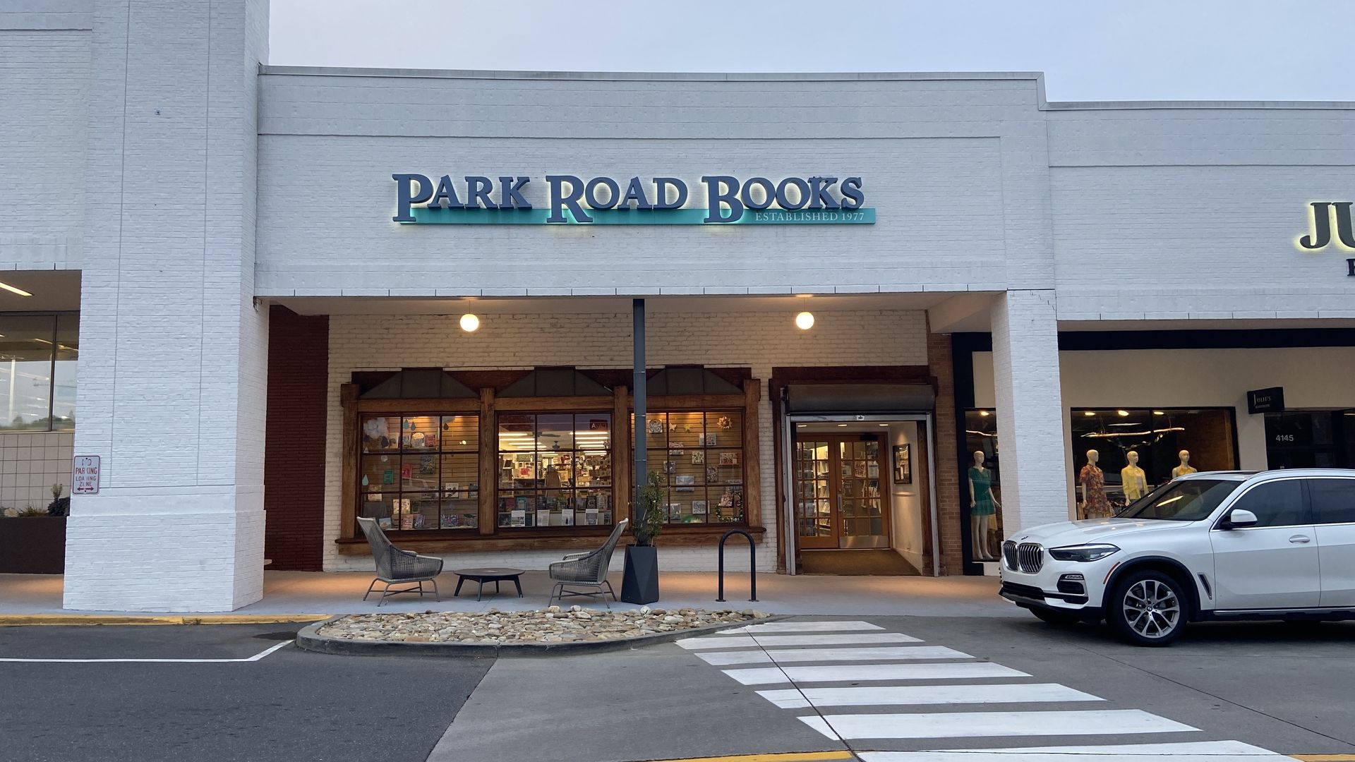 Park Road Books storefront in a white brick strip mall. Large sign reads PARK ROAD BOOKS with a turquoise underline and ESTABLISHED 1977. Glass display windows, main entrance, outdoor chairs, and a white SUV on the right.