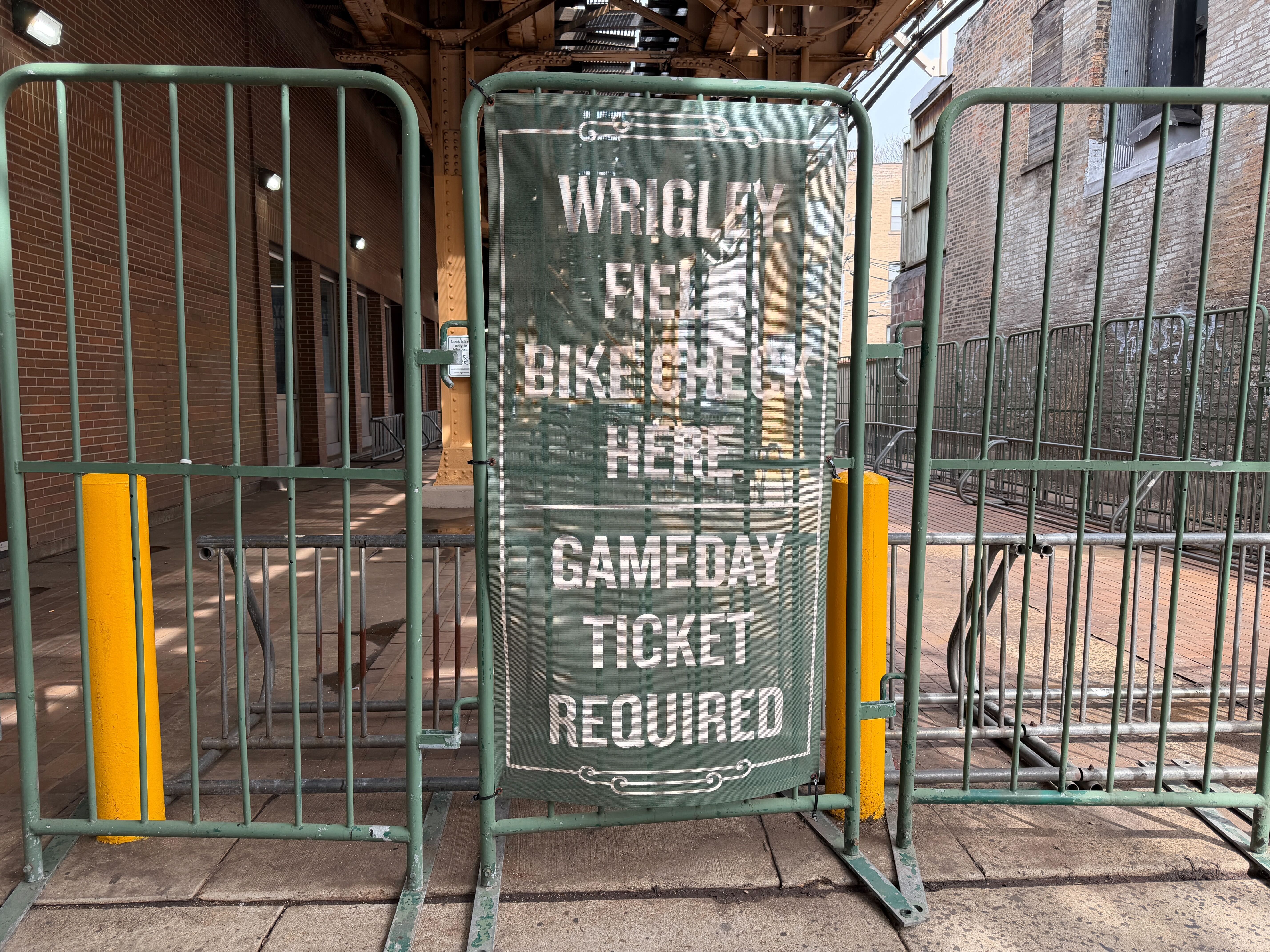 Green stadium gate with a mesh banner reading "WRIGLEY FIELD BIKE CHECK HERE GAMEDAY TICKET REQUIRED" in white letters; brick walls and yellow posts frame the entrance.