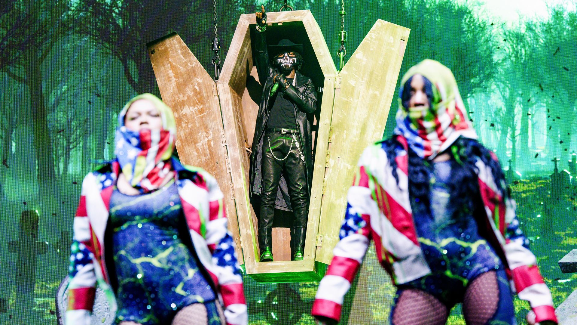 Two backup dancers dance in American flag style jackets in the foreground. In the background but in focus, NBA YoungBoy, wearing all black, sings from the inside of an opened casket.