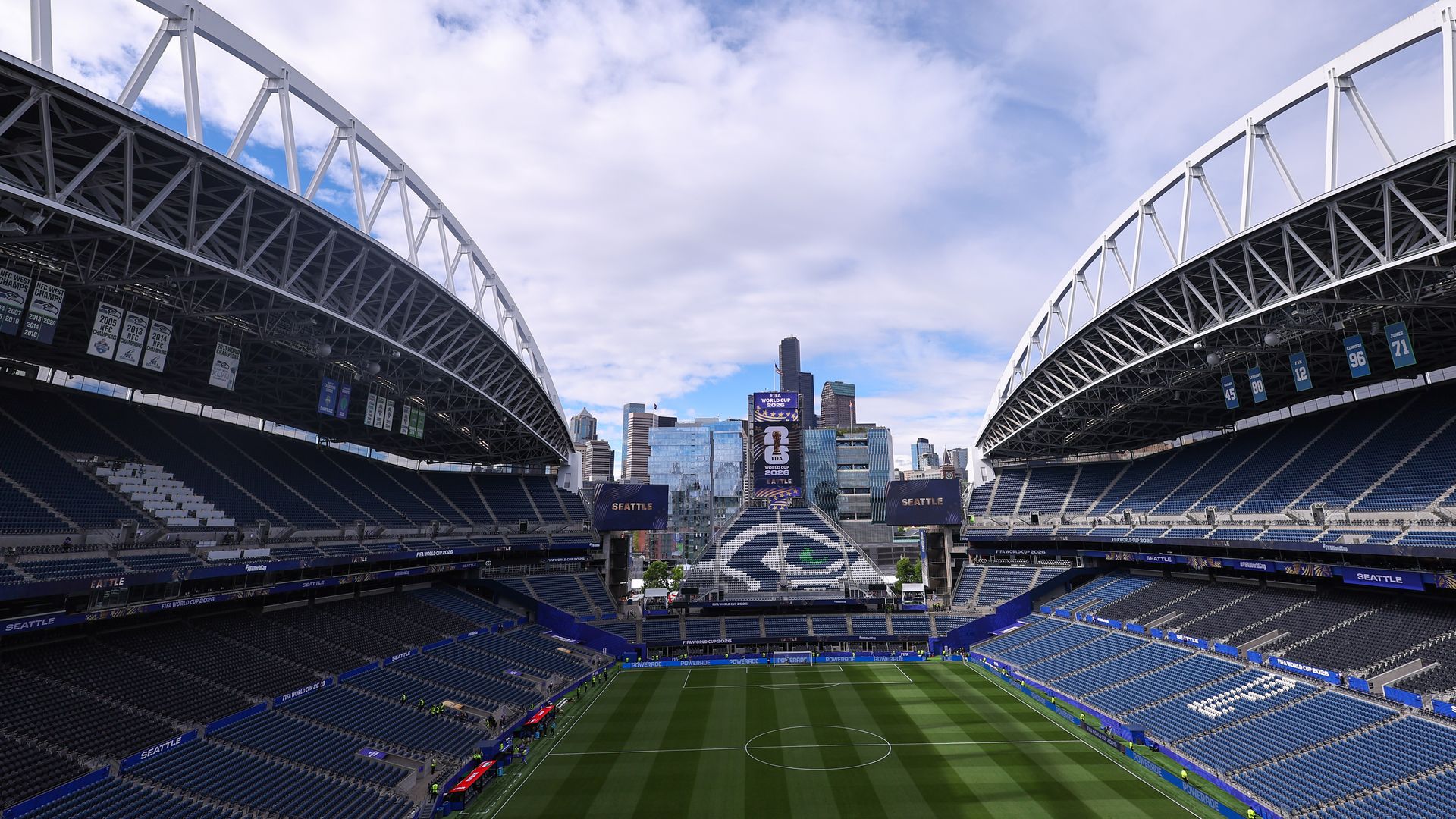 Wide view of a soccer stadium with white steel arches and blue seating, banners along the stands, a green pitch, and a city skyline under a partly cloudy blue sky.