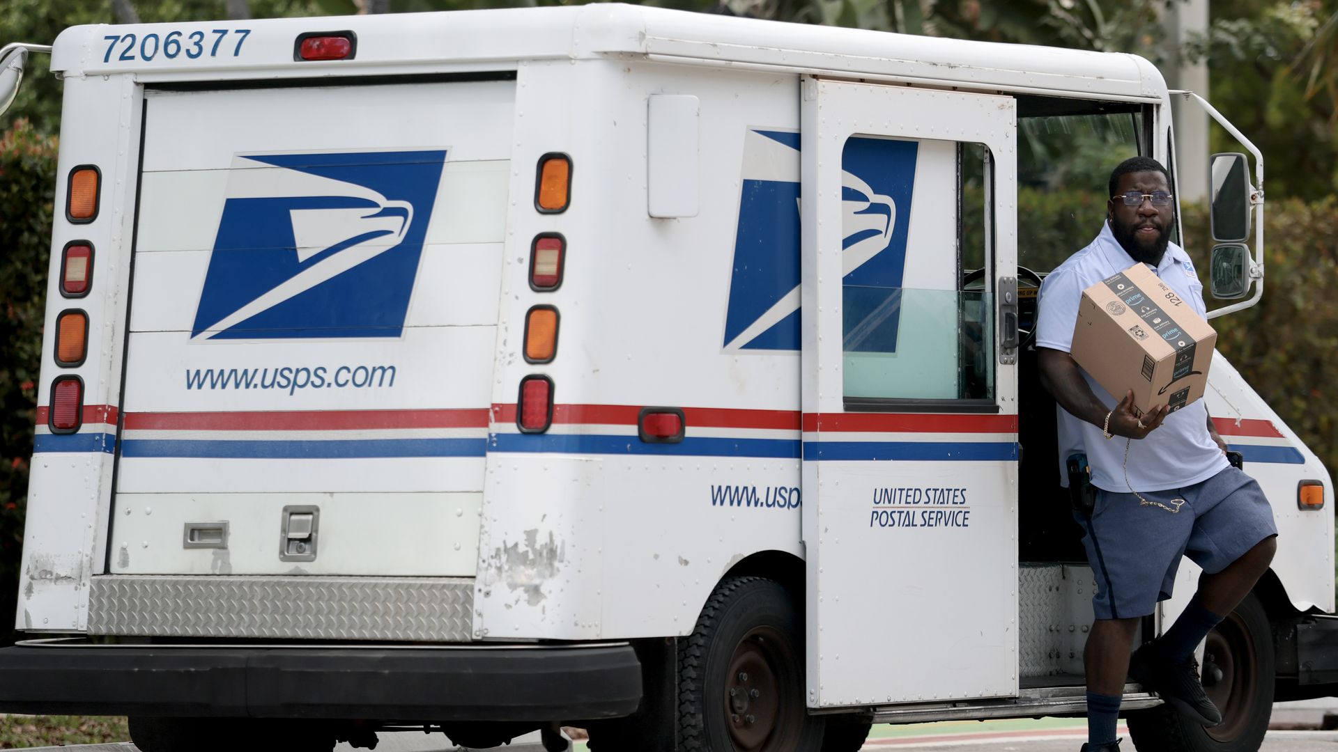 White USPS delivery truck with blue eagle logo and red/blue stripes; a bearded man in a white shirt and blue shorts holds a cardboard box as he steps from the open side door.